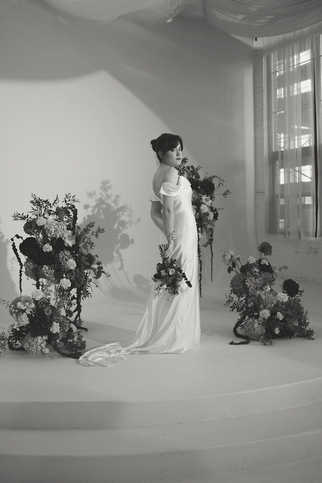 A woman in a satin wedding gown holding a bouquet, standing among floral arrangements in a studio with natural light streaming through a window. marigolds florals nyc florist wedding florals atmosfera wedding
