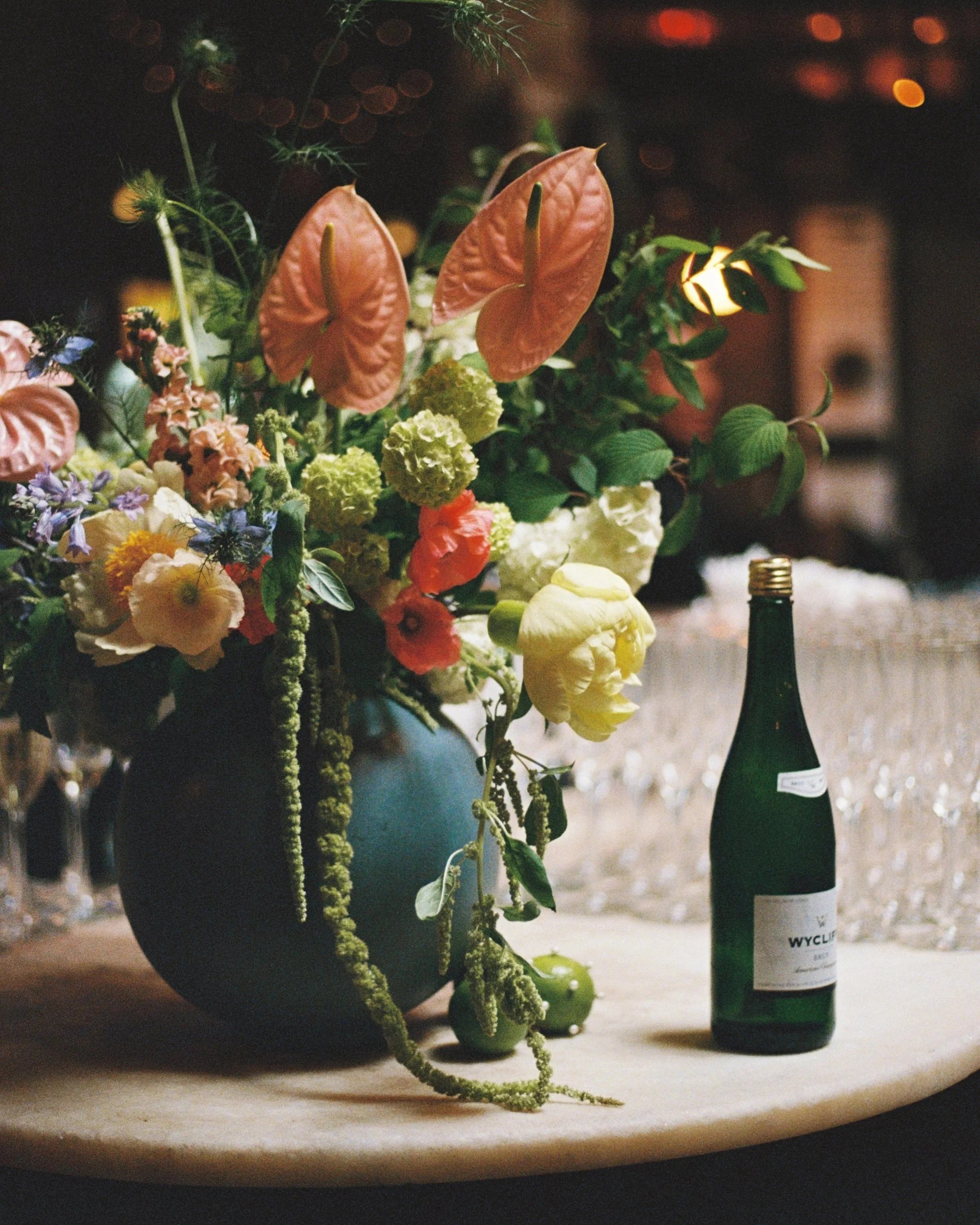 A floral arrangement with pink anthurium flowers, yellow and green hydrangeas, purple and pink smaller flowers, and various greenery in a blue vase, with a green wine bottle on the right side of the image. marigolds florals