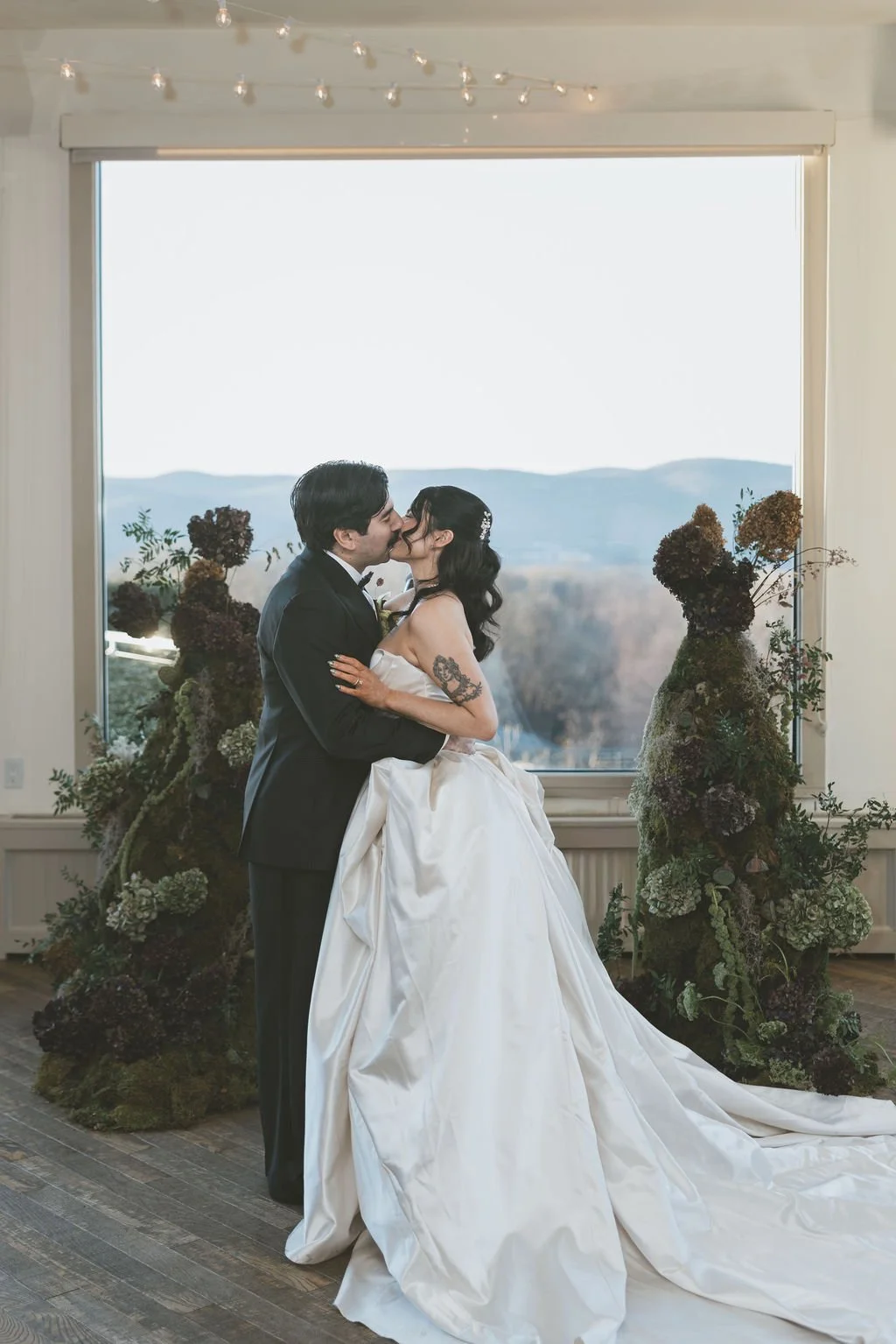 A bride and groom share a kiss during their wedding ceremony, standing by a large window with a mountain view, decorated with floral arrangements.  marigolds florals nyc florist wedding florals the garrison