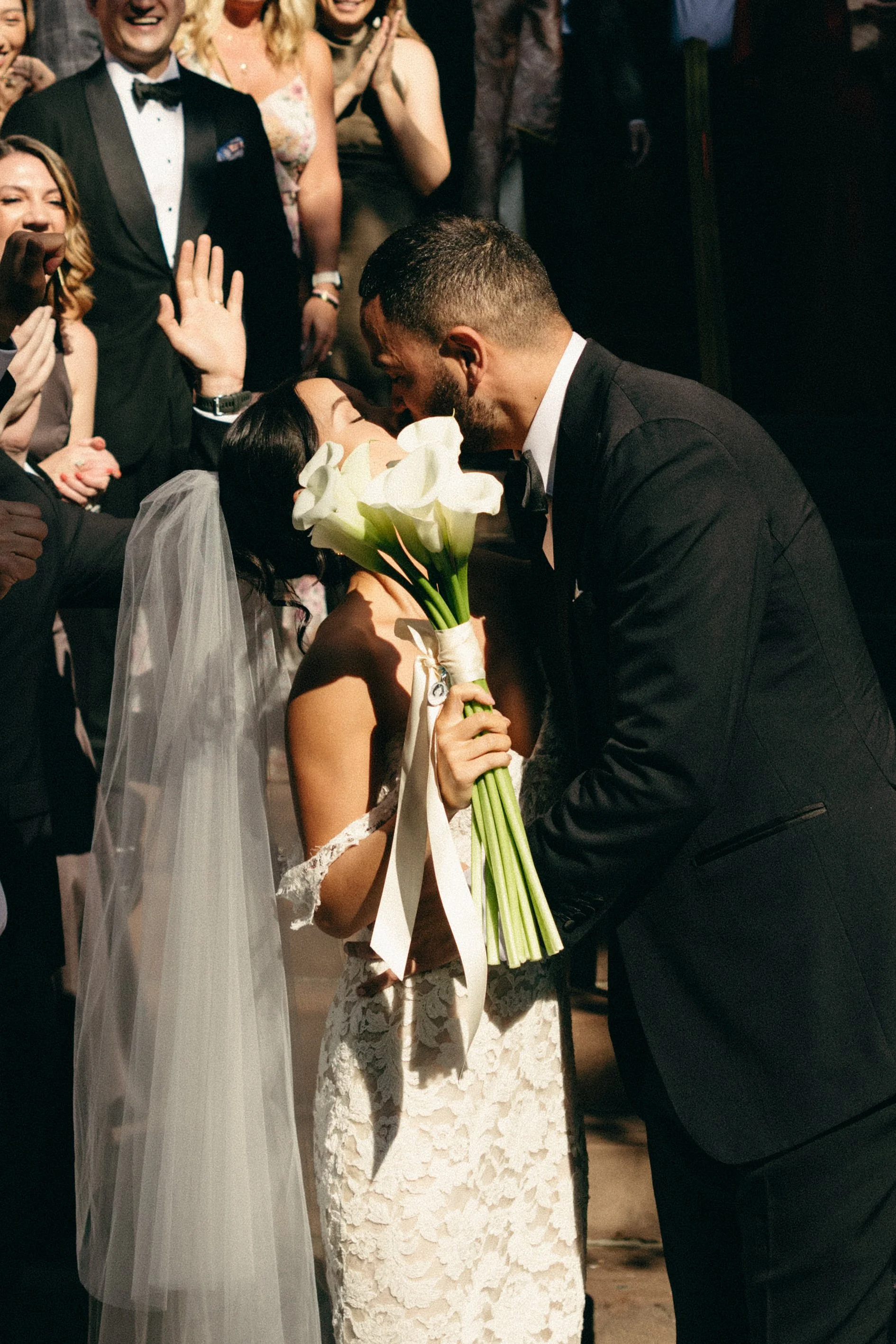 A bride and groom share a kiss during their wedding ceremony, surrounded by seated and standing guests. The bride wears a lace wedding dress and veil, holding a bouquet of white calla lilies, while the groom wears a black tuxedo.  marigolds florals 