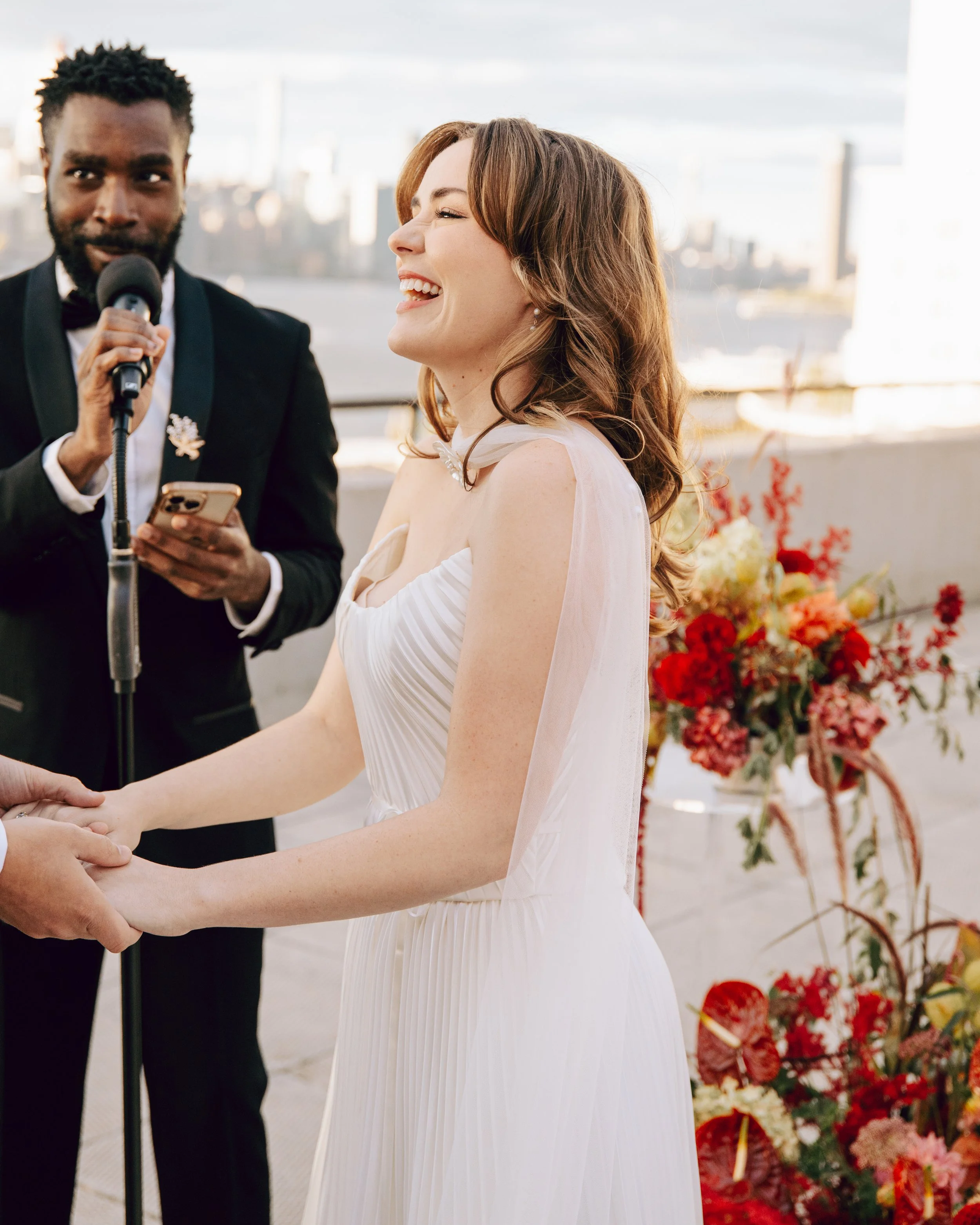 A woman with light skin and brown hair laughing during a wedding ceremony, holding hands with someone. A man in a black tuxedo is speaking into a microphone and looking at her, with a cityscape in the background and a display of flowers behind them.