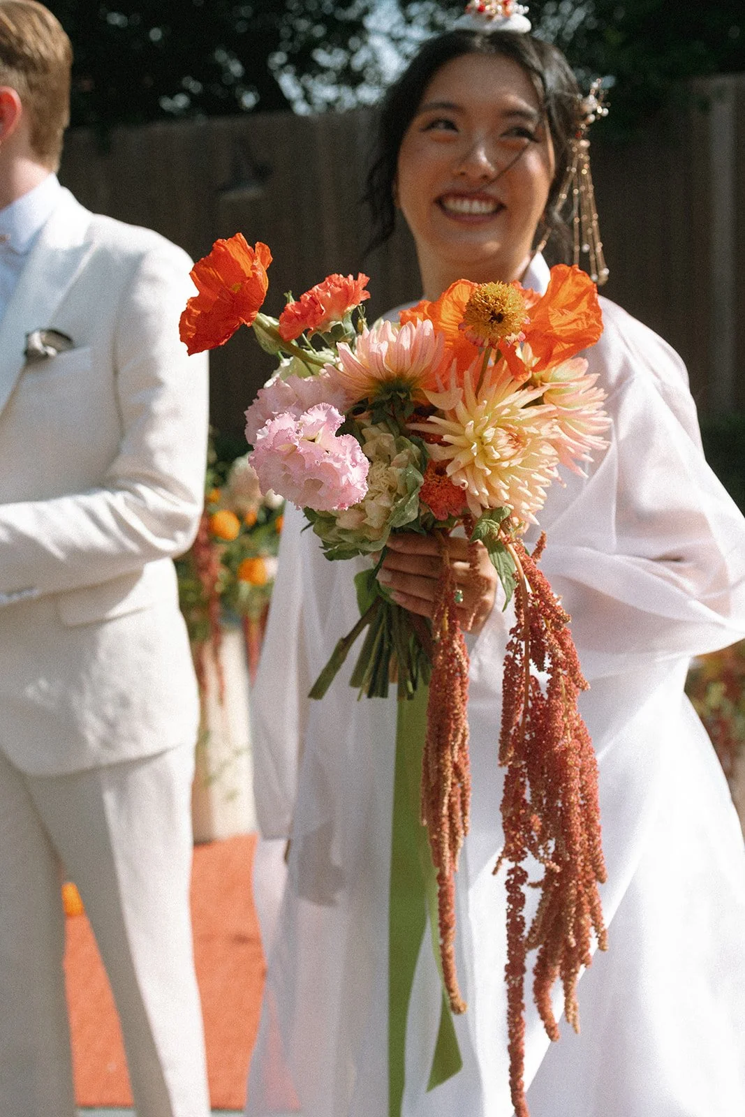 A woman in a white dress smiling while holding a colorful bouquet of flowers, standing outdoors with trees and a wooden fence in the background. marigolds florals nyc florist wedding florals