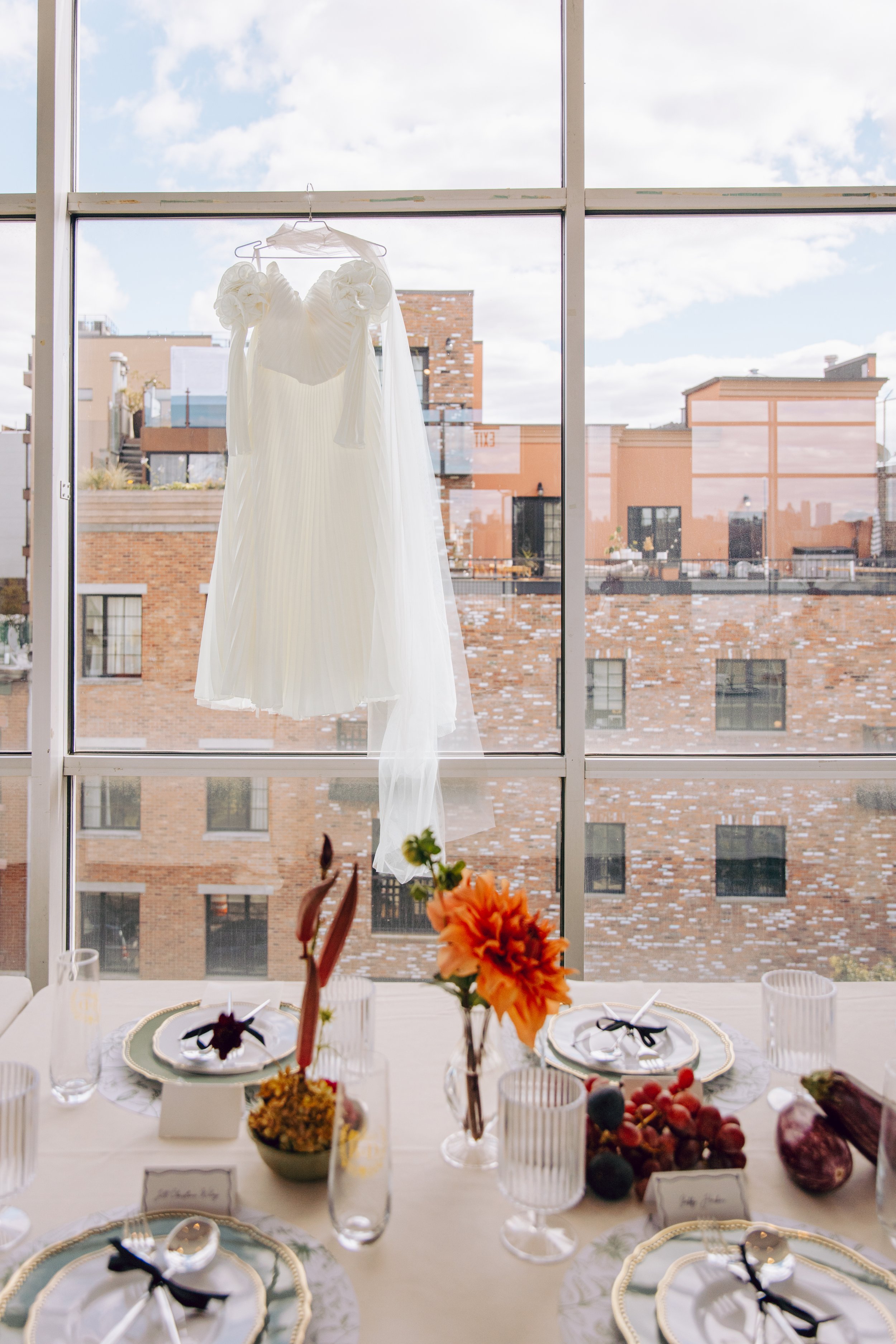 Wedding dress hanging in front of a large window overlooking a cityscape with brick buildings and a blue sky. marigolds florals nyc florist wedding florals the w loft wedding pedestal wedding ceremony anthurium 
