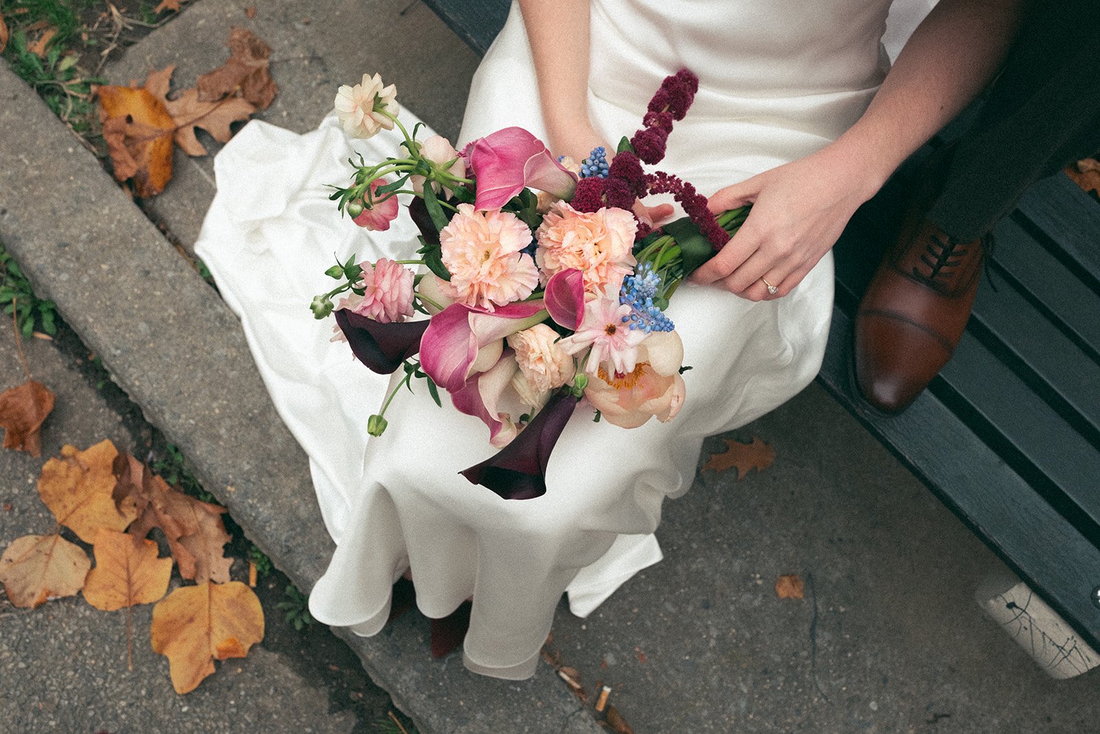 A person in a white dress holding a bouquet of assorted flowers, including calla lilies, carnations, and other blooms, sitting on a park bench with fallen autumn leaves on the ground marigolds florals nyc florist wedding florals atmosfera wedding