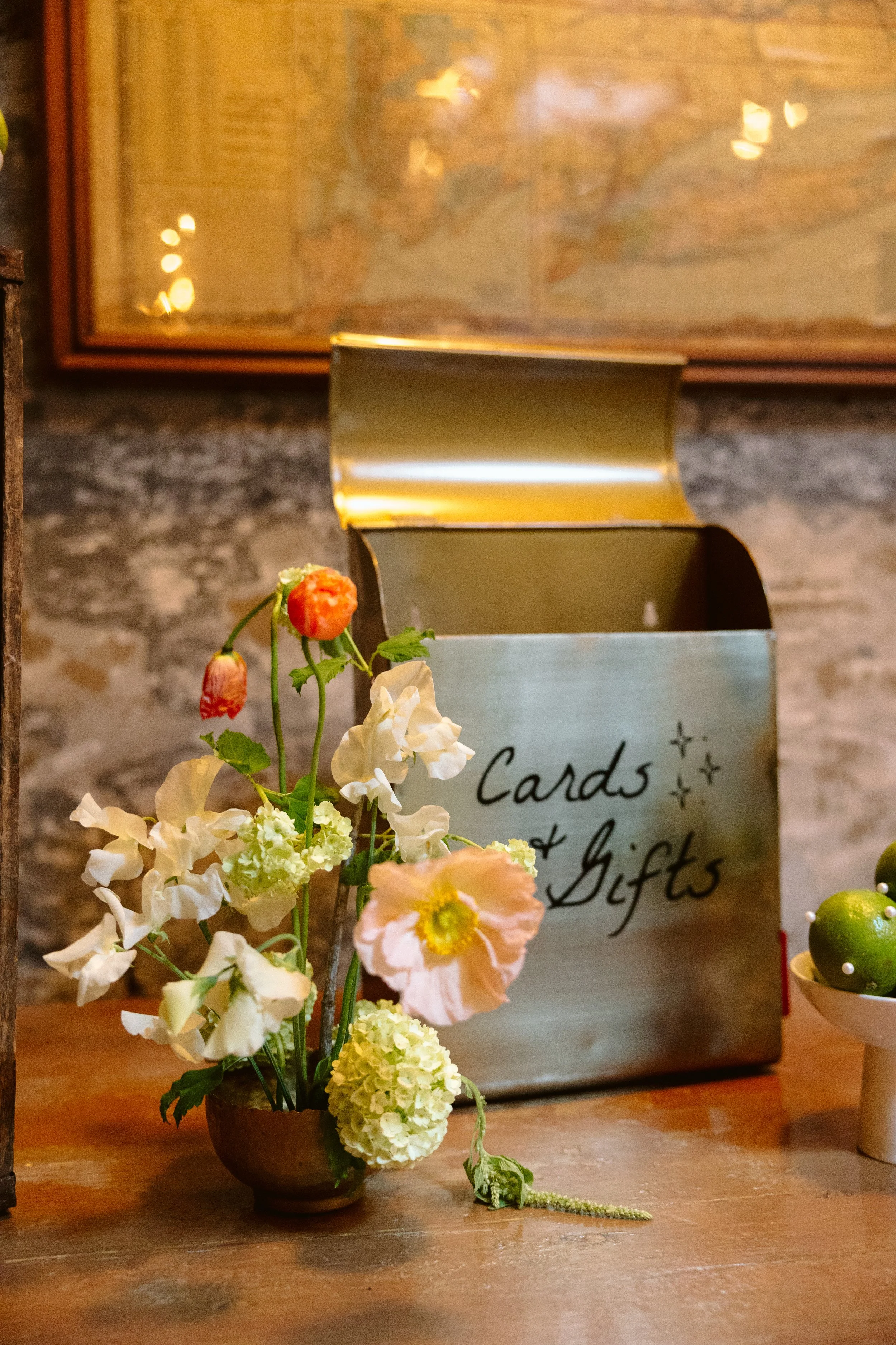 A close-up of a floral arrangement in a brown bowl with white, orange, and pink flowers, placed in front of a metallic container with the partially visible text "Cards + Gifts." The background features a brick wall and a framed map or artwork.