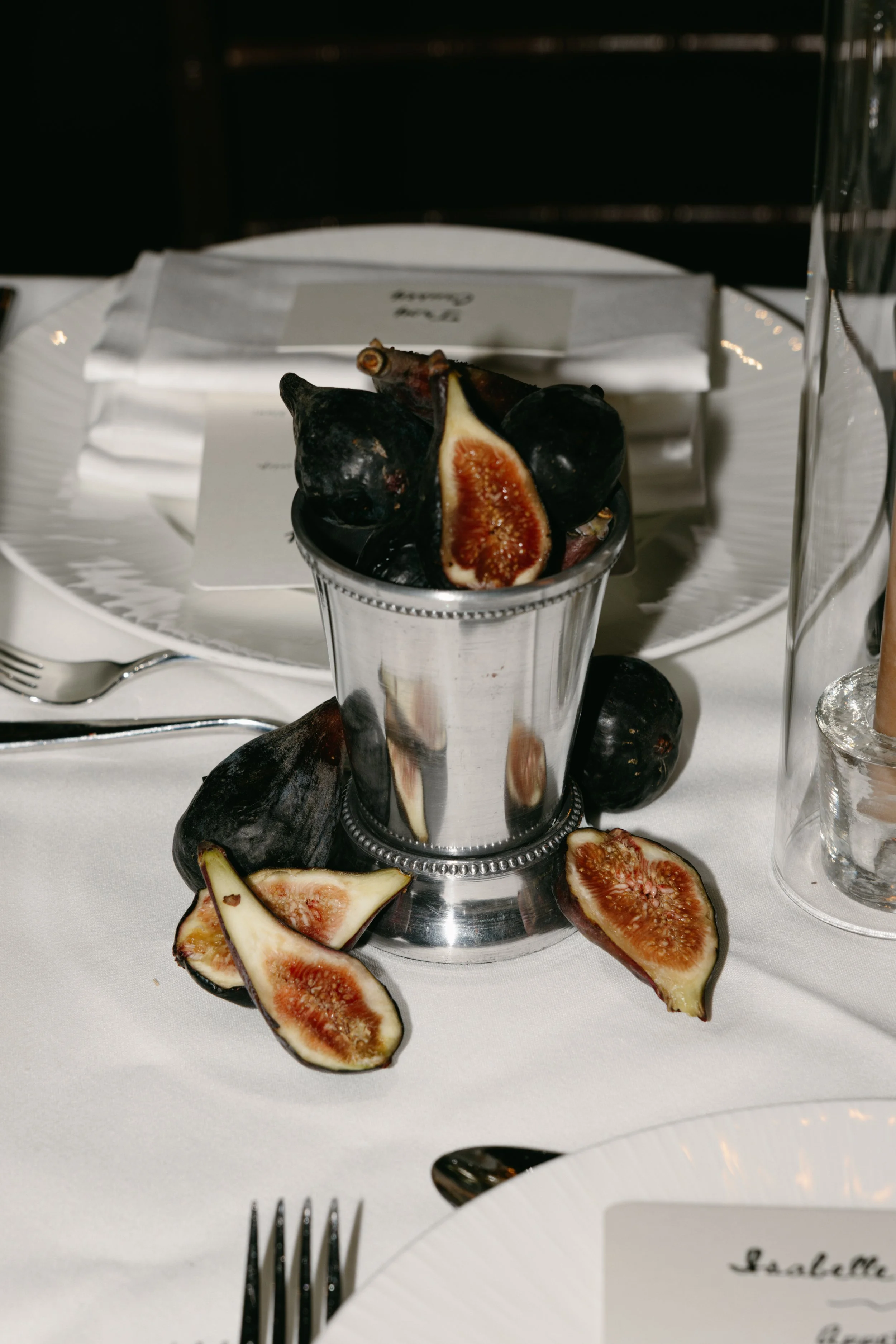 Figs on a table, with some cut open, placed in front of a silver cup, and a white plate with a knife and fork nearby, in a formal dining setting. marigolds florals nyc florist wedding florals central park boathouse wedding