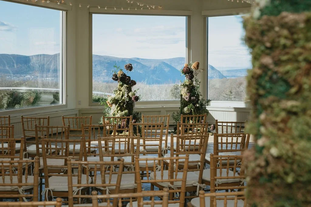 Indoor venue with wooden chairs arranged for a wedding or event, large windows showing mountains and blue sky, floral arrangements, and string lights overhead. marigolds florals nyc florist wedding florals atmosfera wedding