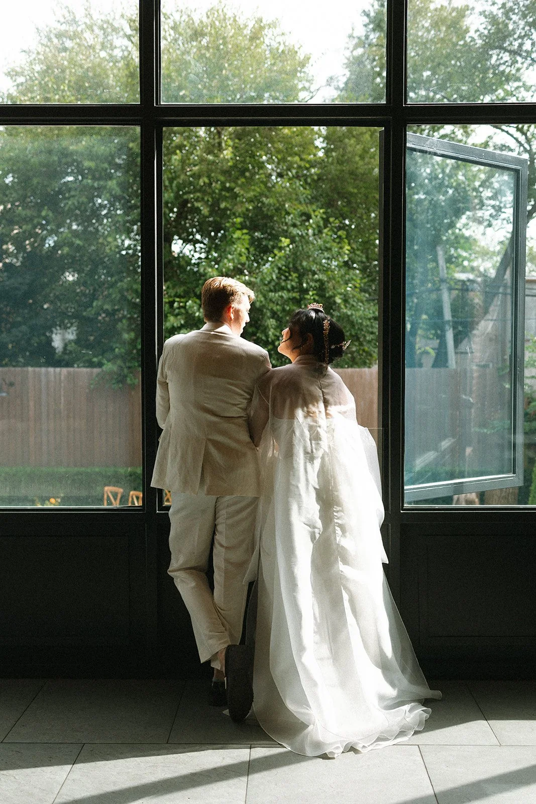 A couple dressed in wedding attire standing by an open window, looking outside at green trees. marigolds florals nyc florist wedding florals