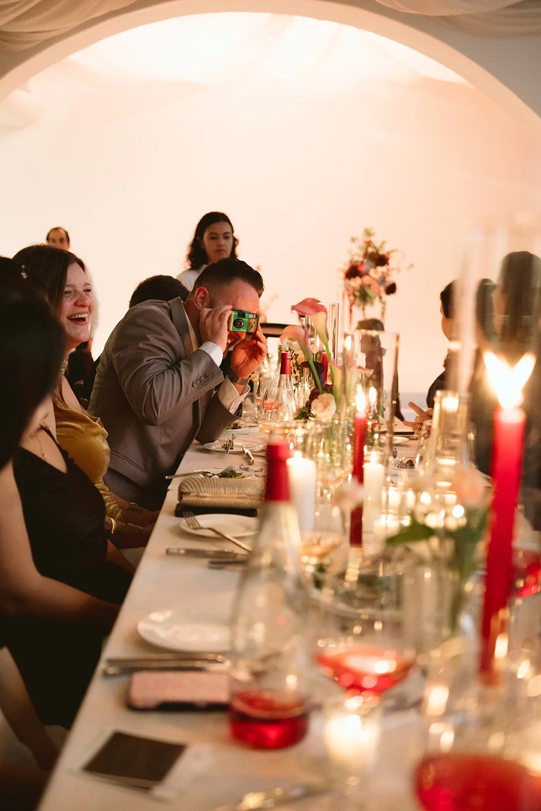People sitting at a decorated banquet table during a celebration, with candles, flowers, and guests taking photos. marigolds florals nyc florist wedding florals atmosfera wedding intimate wedding
