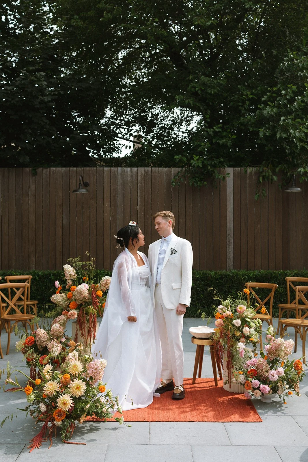 A couple in wedding attire standing on an outdoor red rug, surrounded by floral arrangements, with wooden chairs and a wooden fence in the background. marigolds florals