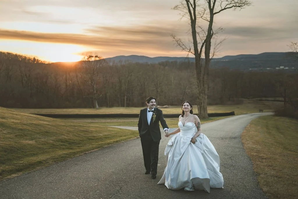 A newlywed couple walking hand in hand on a winding path during sunset, with a scenic landscape and bare trees in the background.  marigolds florals nyc florist wedding florals the garrison