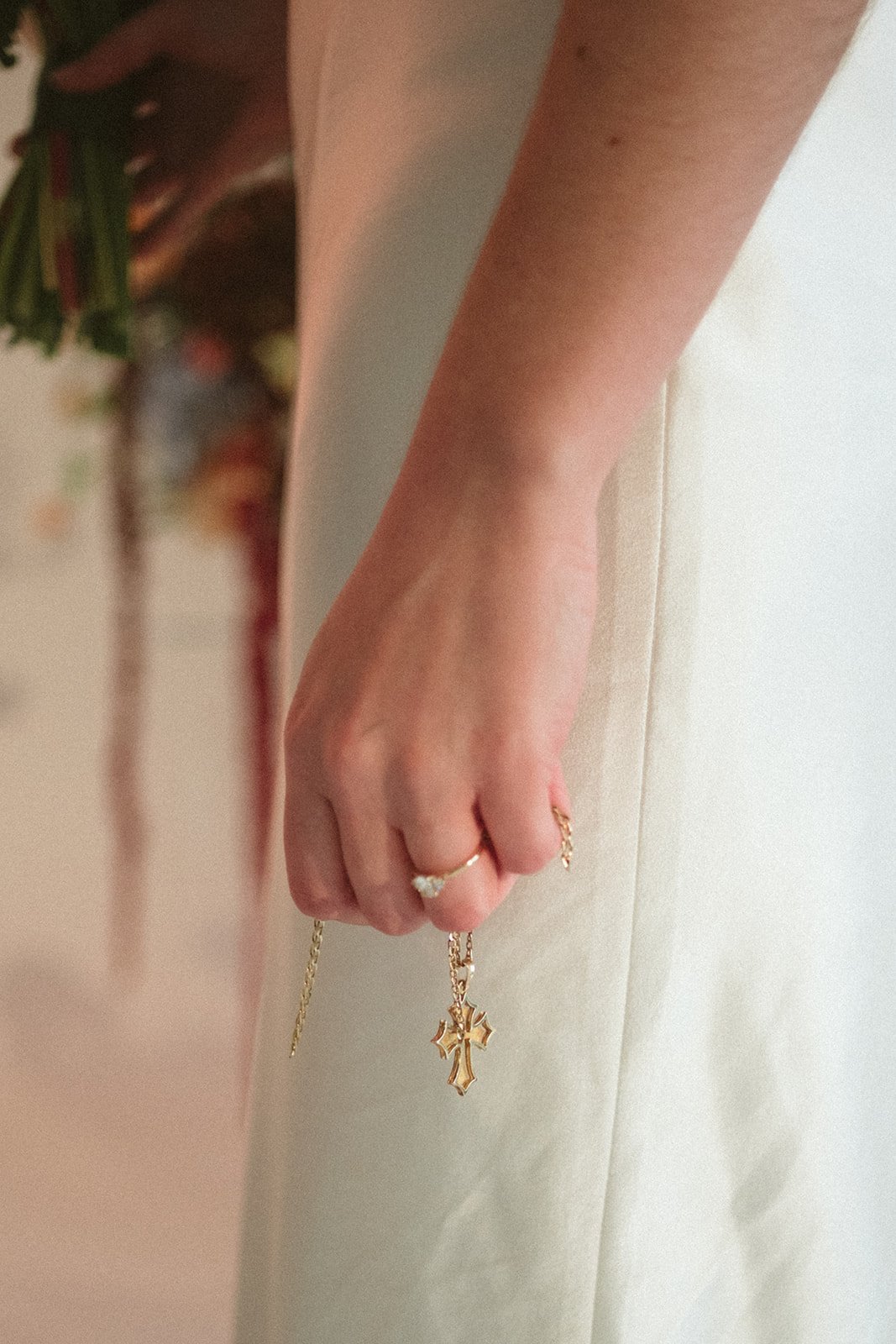 Close-up of a hand holding a gold cross necklace, with rings on fingers, against a beige background. marigolds florals nyc florist wedding florals atmosfera wedding