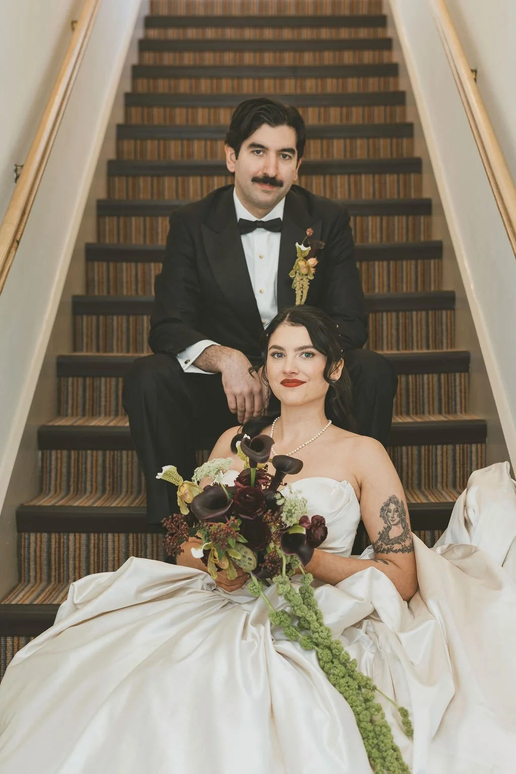 A newlywed couple sitting on a staircase. The groom is in a black tuxedo with a bow tie, and the bride is in a white wedding dress holding a dark floral bouquet.  marigolds florals nyc florist wedding florals the garrison