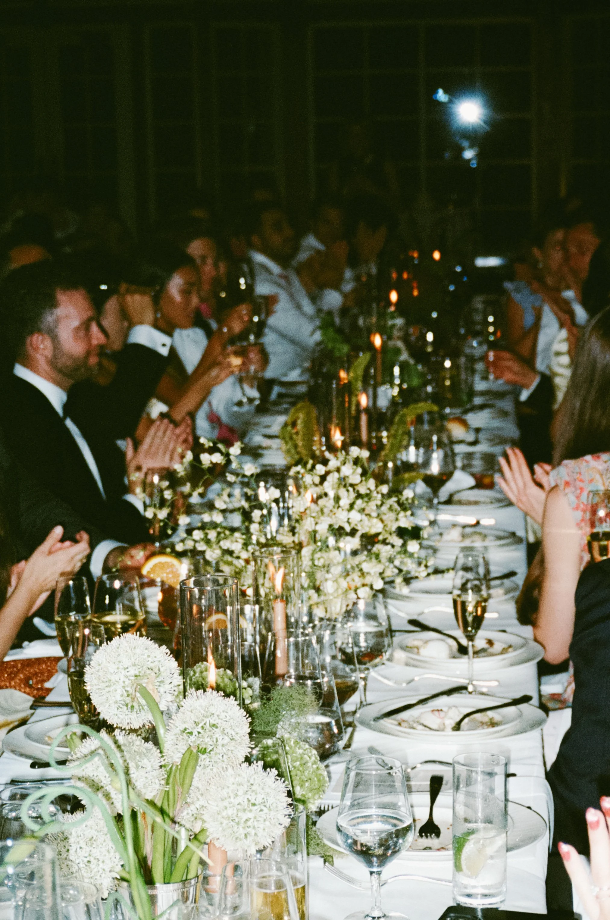 People sitting at a long dinner table with floral centerpieces, glasses of wine and candles, in a dimly lit setting. marigolds florals nyc florist wedding florals central park boathouse wedding