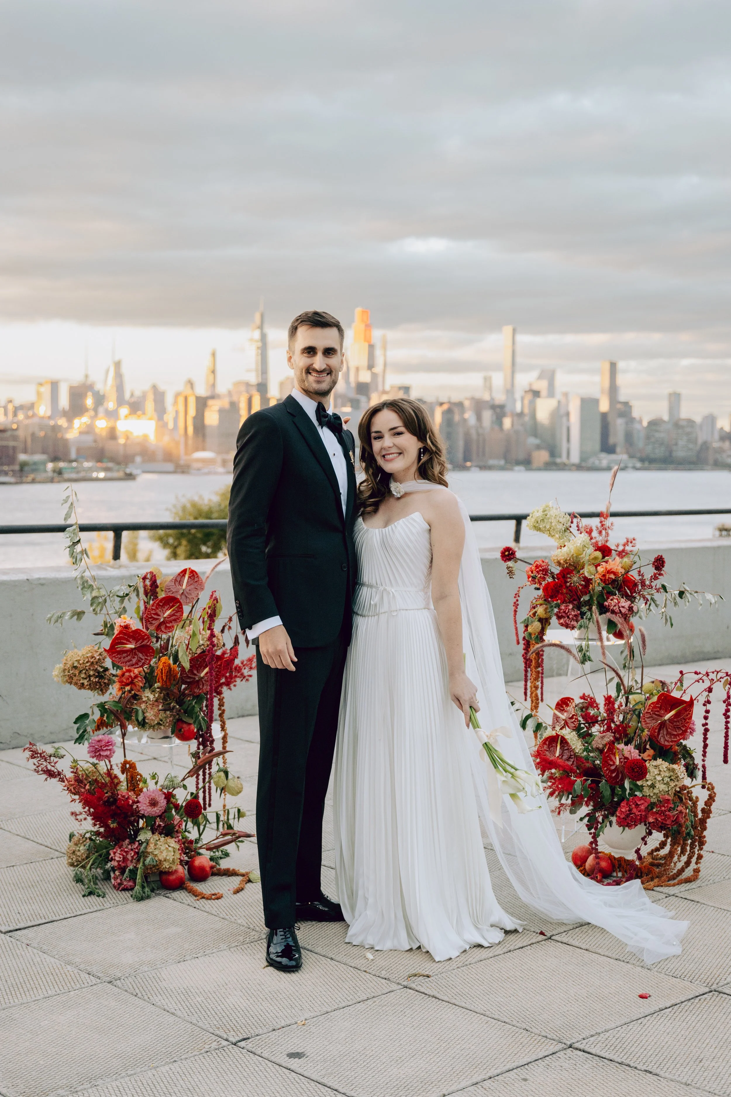 A newlywed couple in wedding attire standing outdoors with a city skyline in the background, decorated with floral arrangements featuring red and pink flowers and pomegranates. marigolds florals nyc florist wedding florals the w loft wedding pedestal