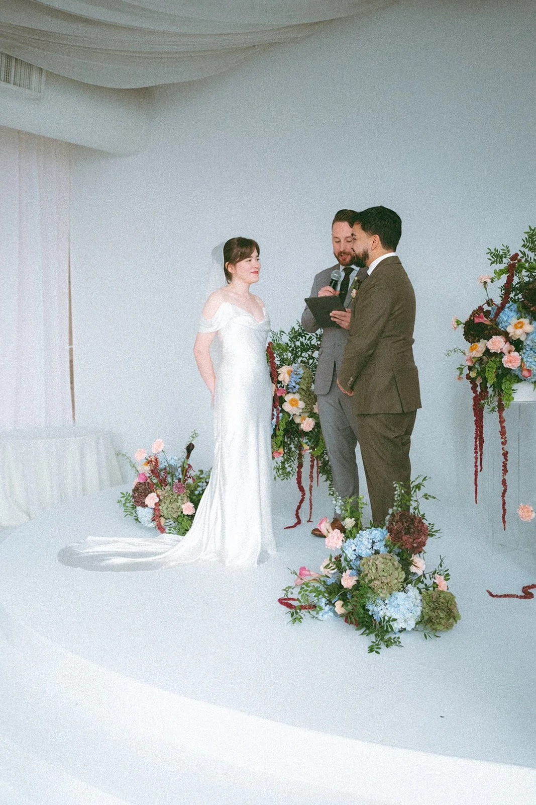 A wedding ceremony with a bride and groom standing in front of officiant, surrounded by floral arrangements, on a white backdrop. marigolds florals nyc florist wedding florals atmosfera wedding marigolds florals nyc florist wedding florals atmosfera 