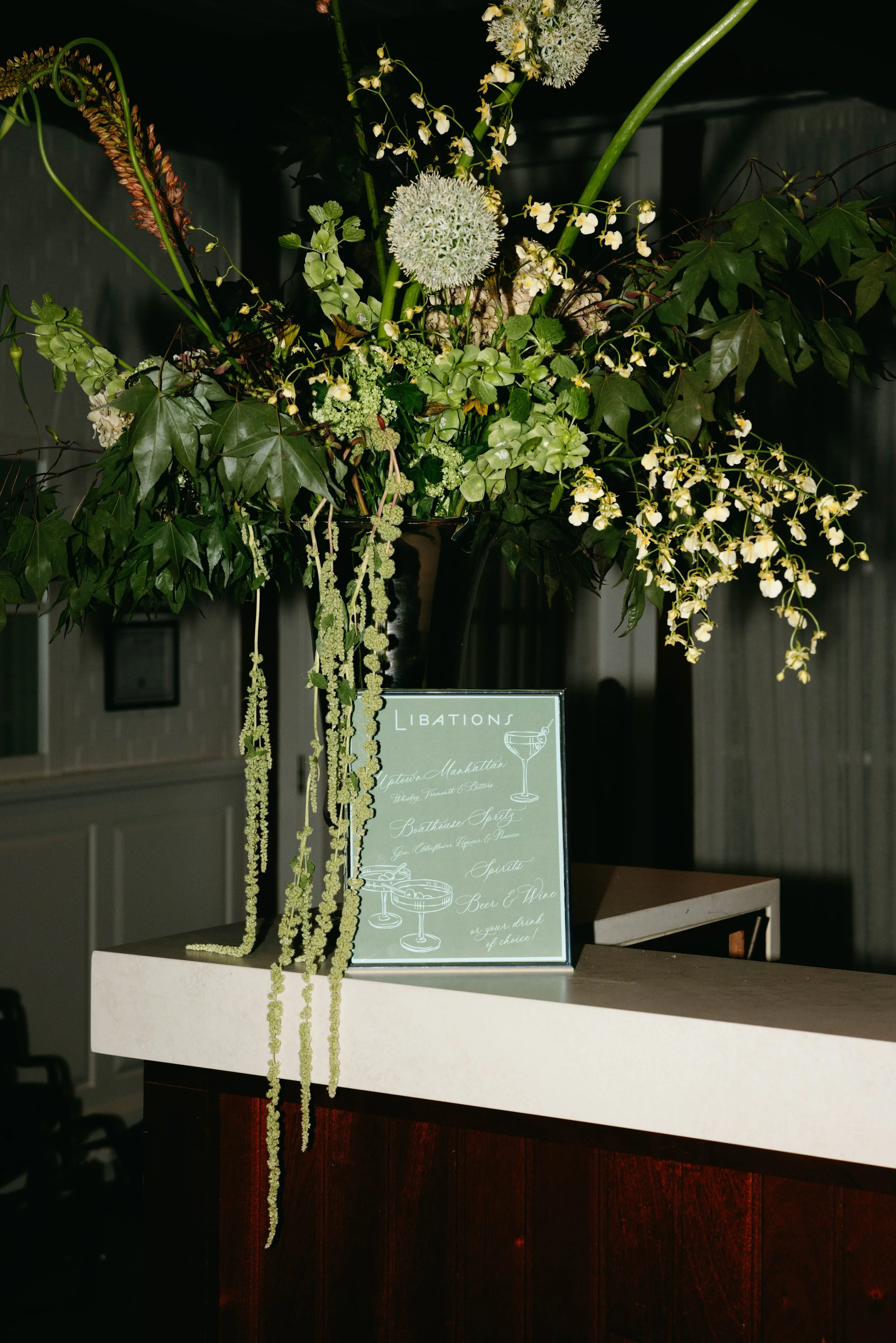 A floral arrangement with white and green flowers and large green leaves on a white counter, with a light-colored club-themed menu sign in front. marigolds florals nyc florist wedding florals central park boathouse wedding