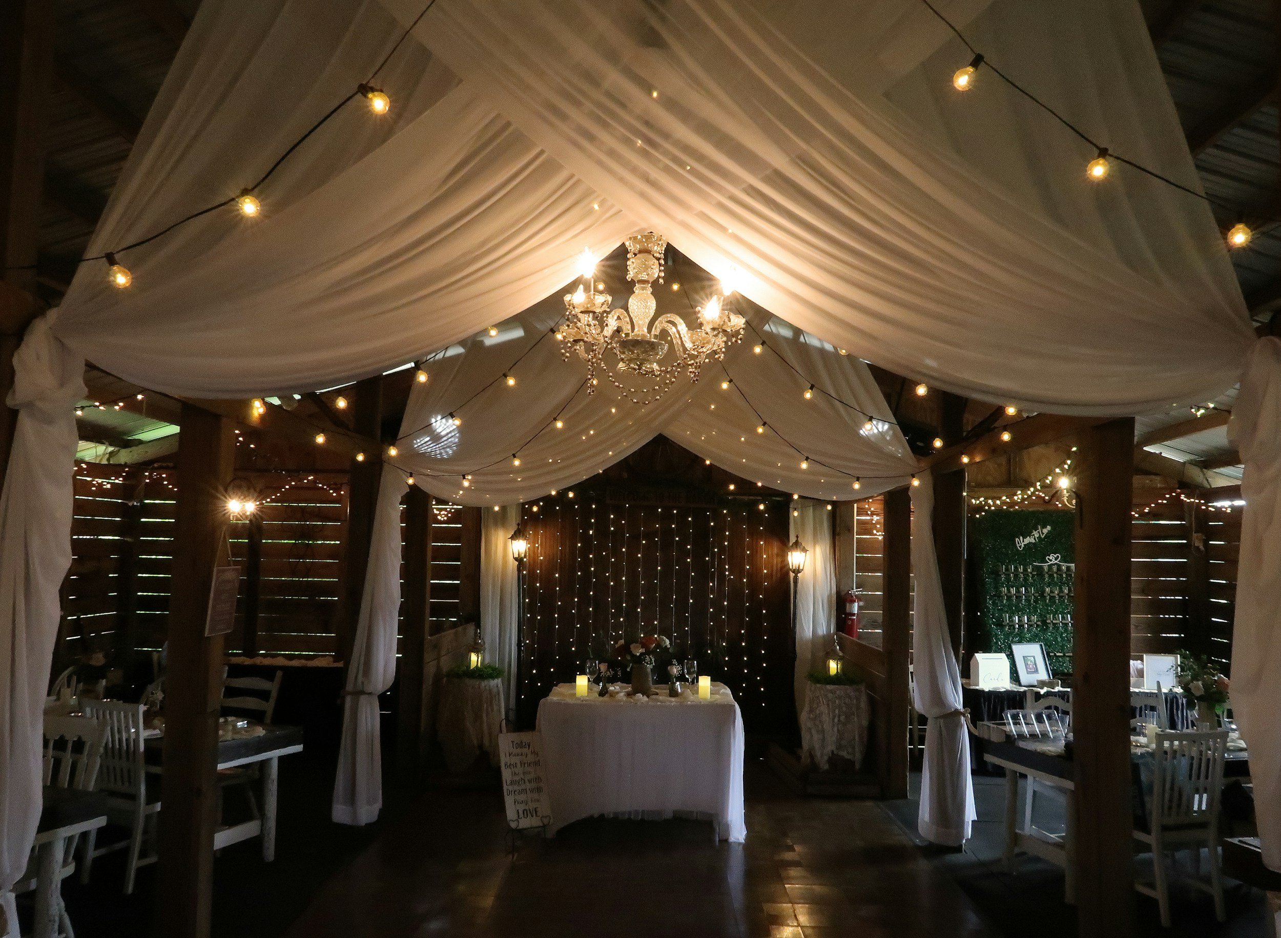 Decorated event space with draped white fabric ceiling, string lights, chandelier, tables, and chairs.