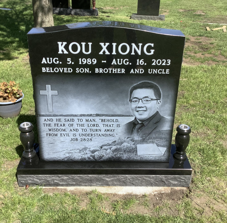 Gravestone for Kou Xiong, who lived from August 5, 1989, to August 16, 2023. It is located in a cemetery with grass, trees, and other graves nearby. The gravestone features a cross and an engraved photo of Kou Xiong in glasses and a suit. It also includes a Bible verse from Job 28:28.