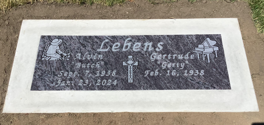 A grave marker with the surname 'Lebens,' featuring images of a girl planting flowers, a cross, and a grand piano, and the names and birth and death dates of Lucy Butch and Gertrude Gerty.