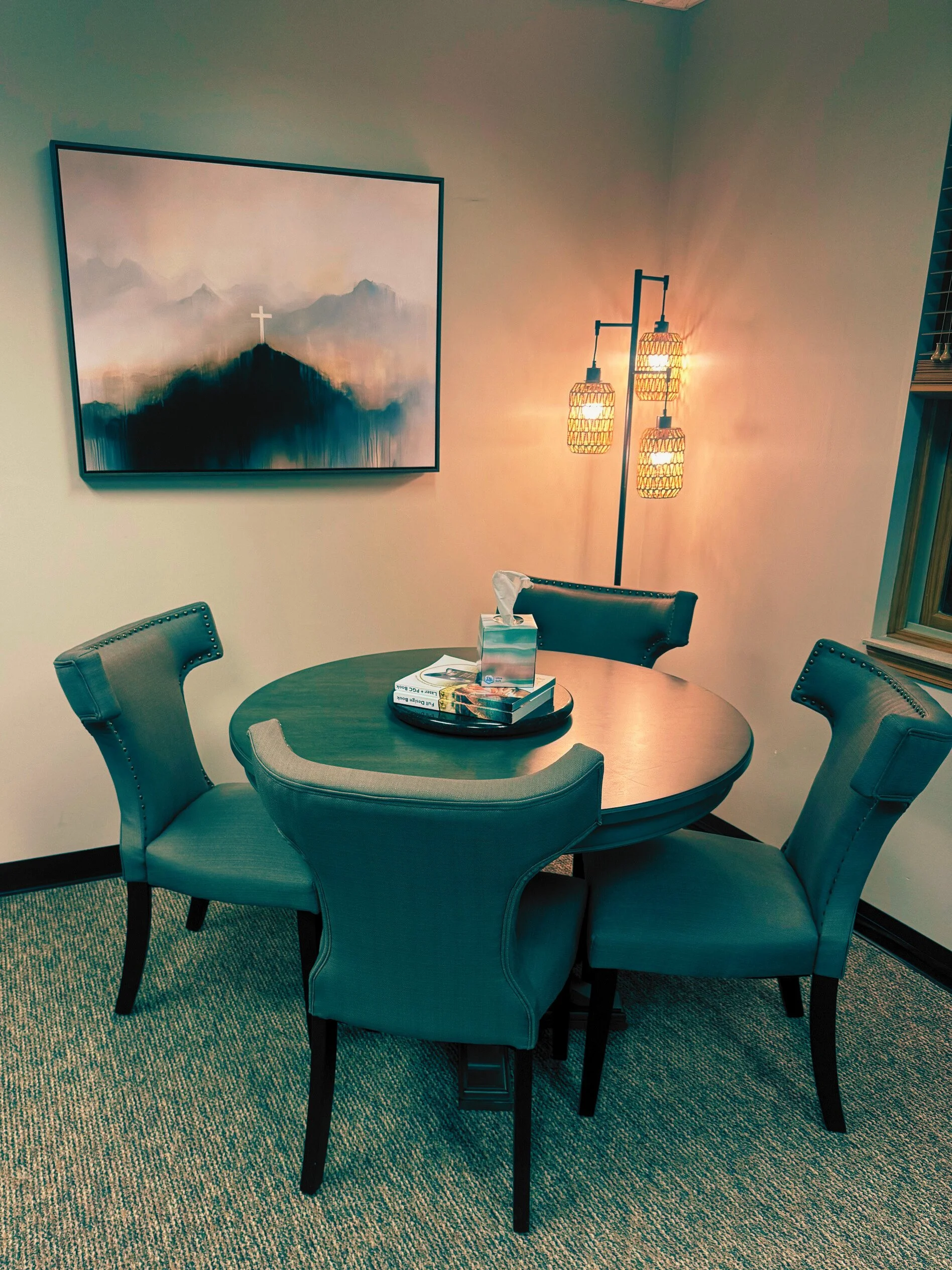 A dining room with a round table surrounded by five green upholstered chairs, a tray with books and a tissue box on the table, a wall-mounted painting of a mountain with a cross, and a floor lamp with three wicker shades emitting warm light.