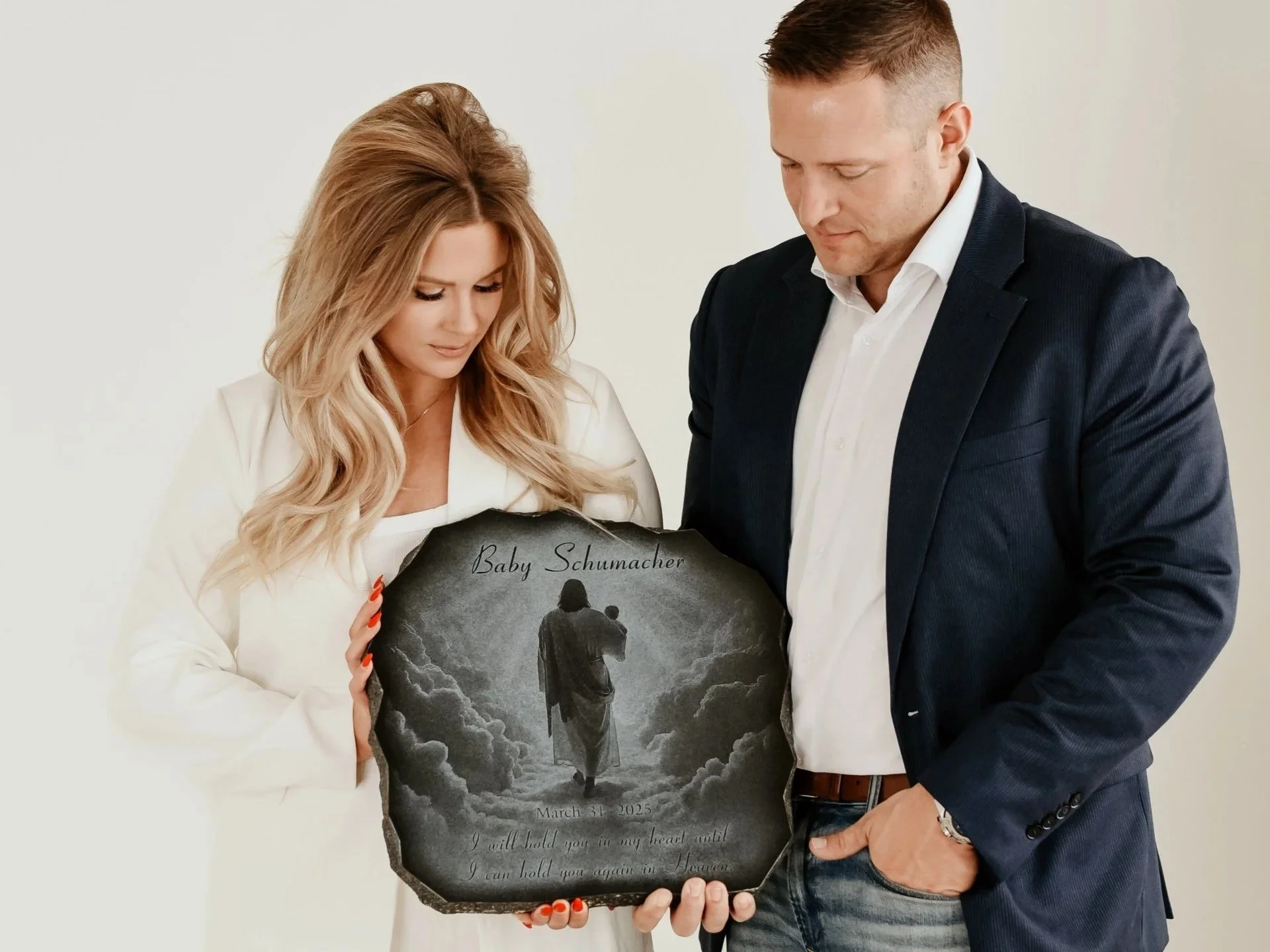 A woman with long blonde hair and a man with short brown hair looking down at a black memorial plaque with a picture of a person walking away in the clouds and the words 'Baby Schumacher,' a date, and a message about holding someone in their heart.