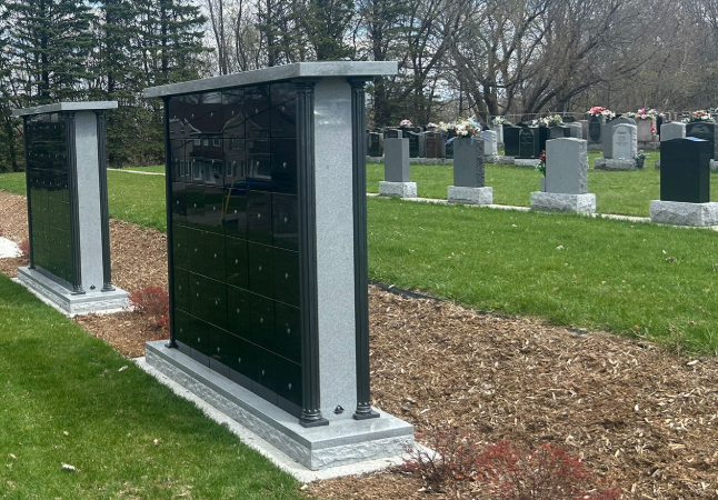 Two dark gray plaques with columns on a grassy area in a cemetery, with other gravestones and trees in the background.