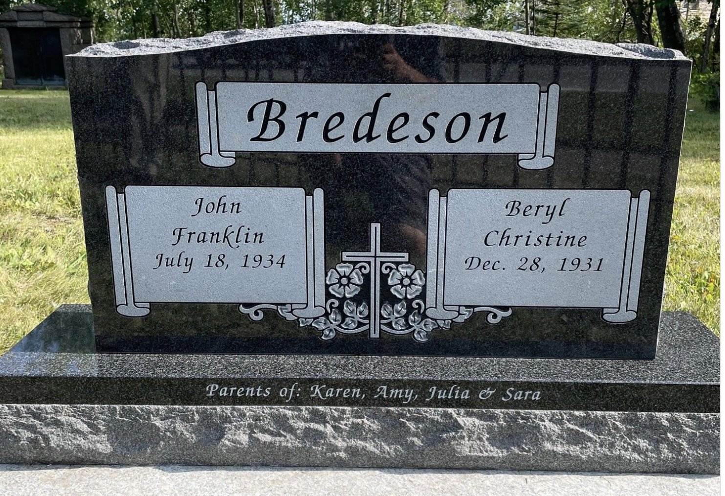 Black granite headstone with engraved silver lettering for the Bredeson family, featuring names John Franklin (born July 18, 1934) and Beryl Christine (born Dec. 28, 1931), includes a Christian cross with floral decoration, and a line of smaller text at the bottom identifying the parents as Karen, Amy, Julia, and Sara.
