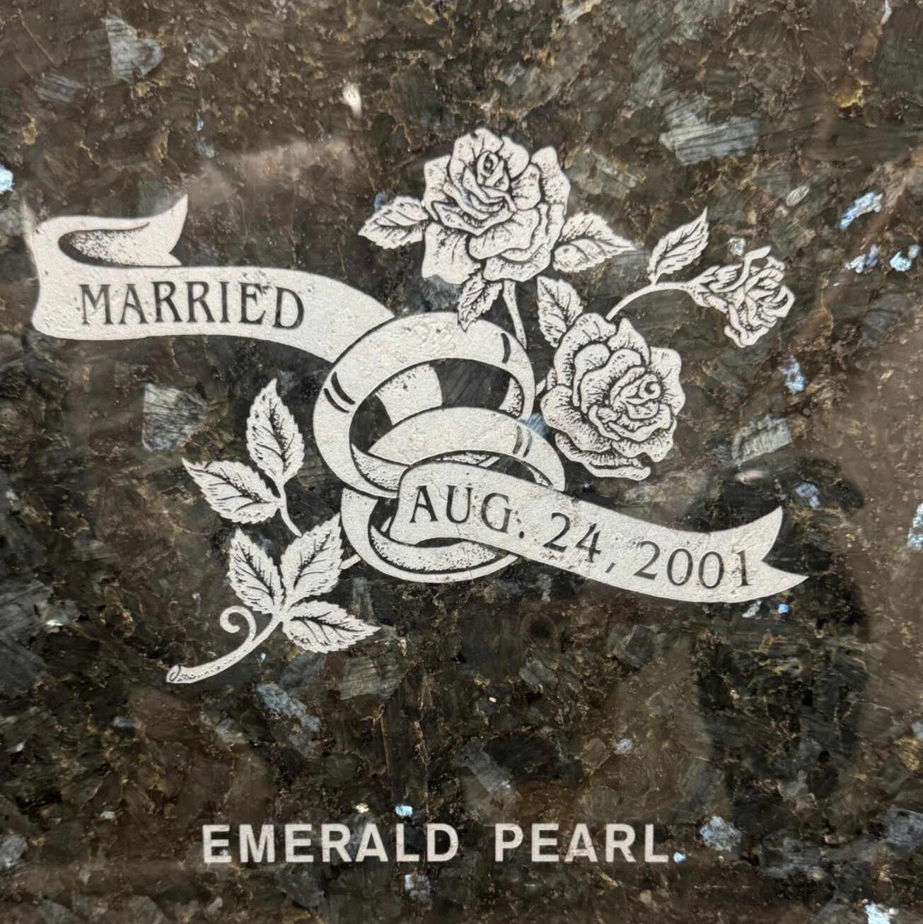A gravestone or memorial plaque with engraved roses and wedding rings symbolizing a marriage on August 24, 2001, with the words 'Married' and 'Emerald Pearl'.