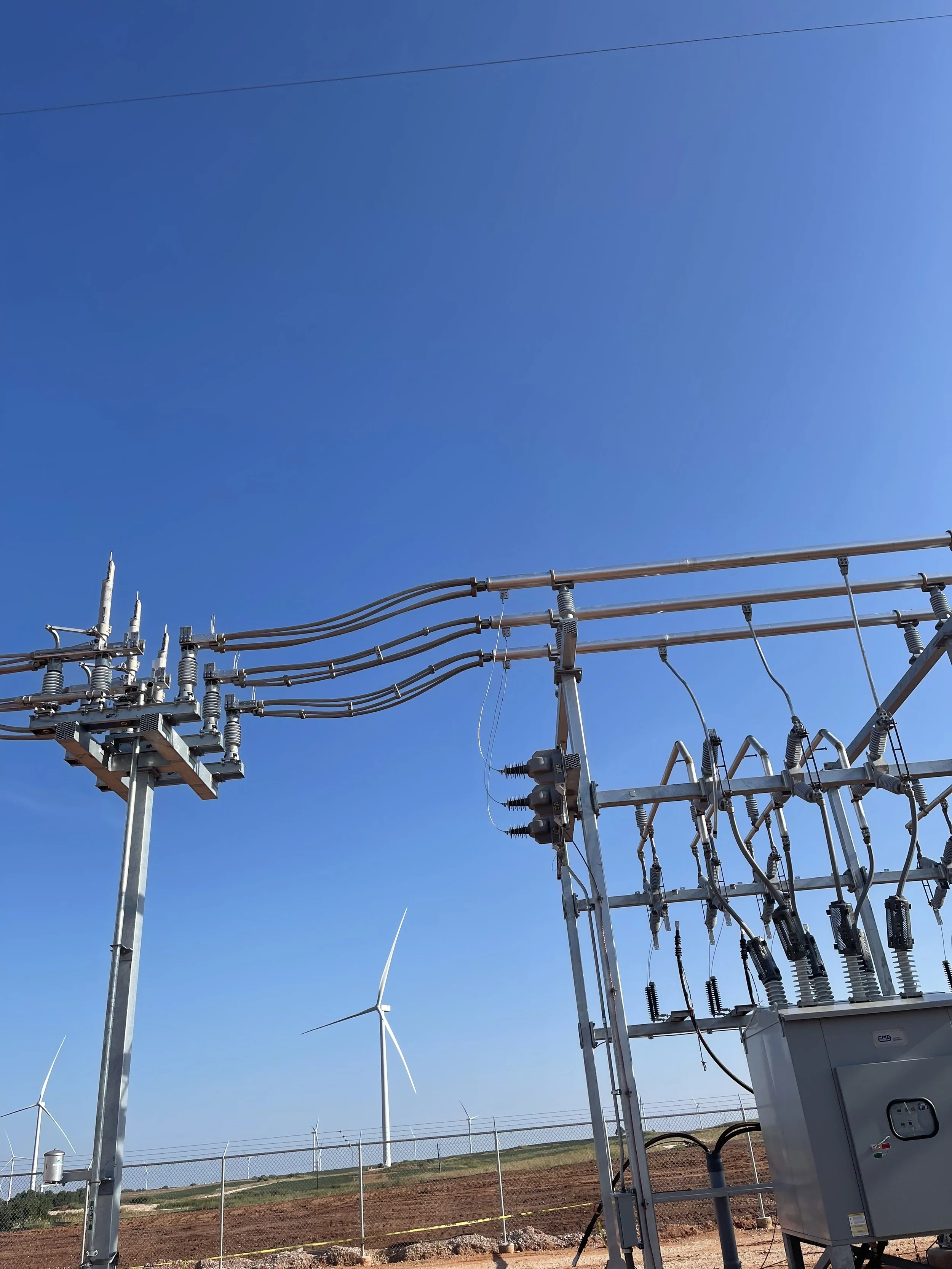 Electrical power lines and equipment with wind turbines in a rural area under a blue sky.