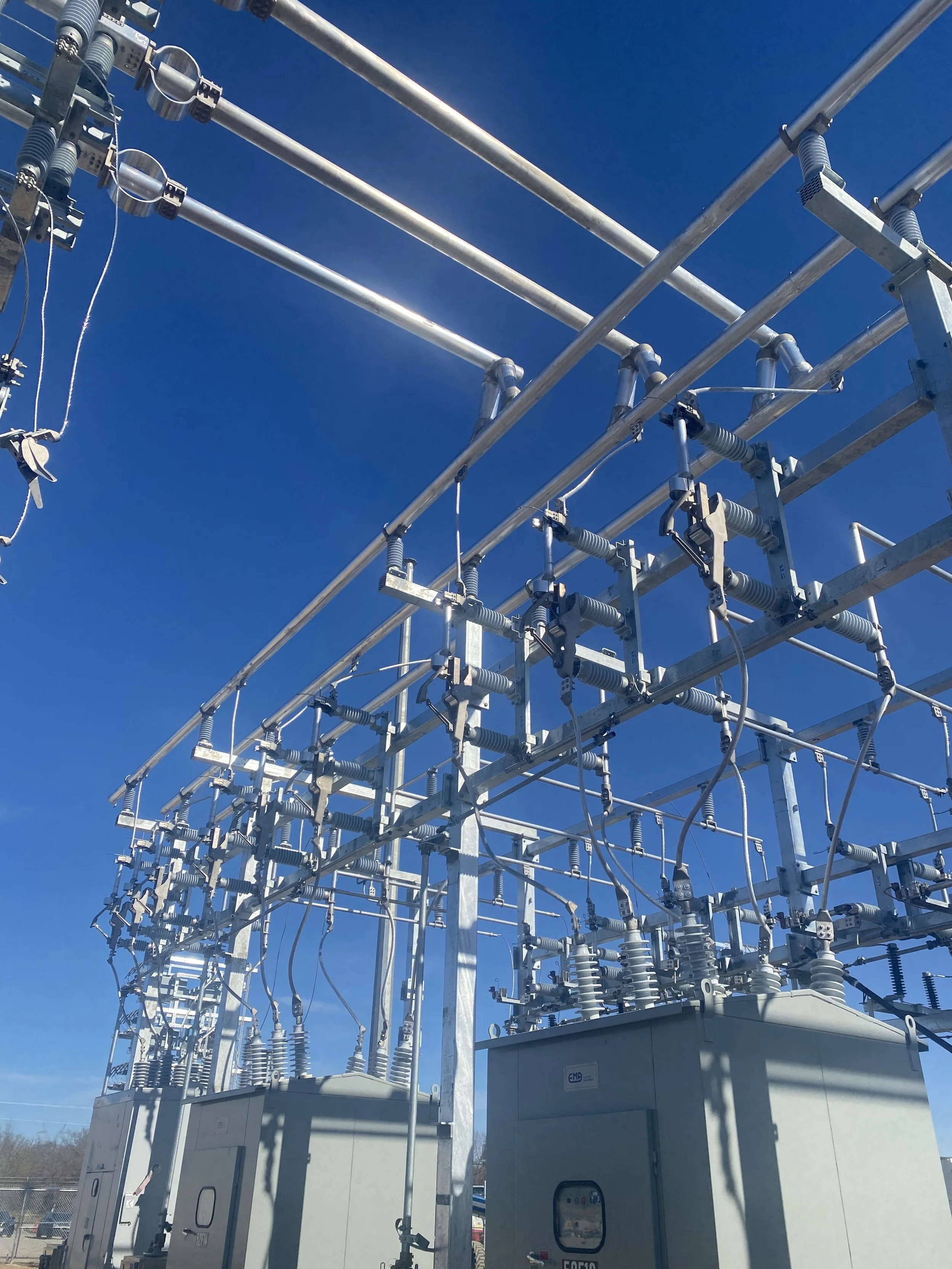 Electrical utility substation with metal structures, circuit breakers, insulators, and transformers against a clear blue sky.