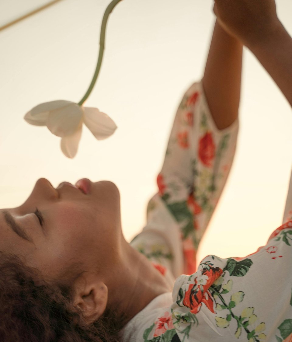 Person lying down with eyes closed, holding a white flower above their face, wearing a floral shirt, with a warm background.