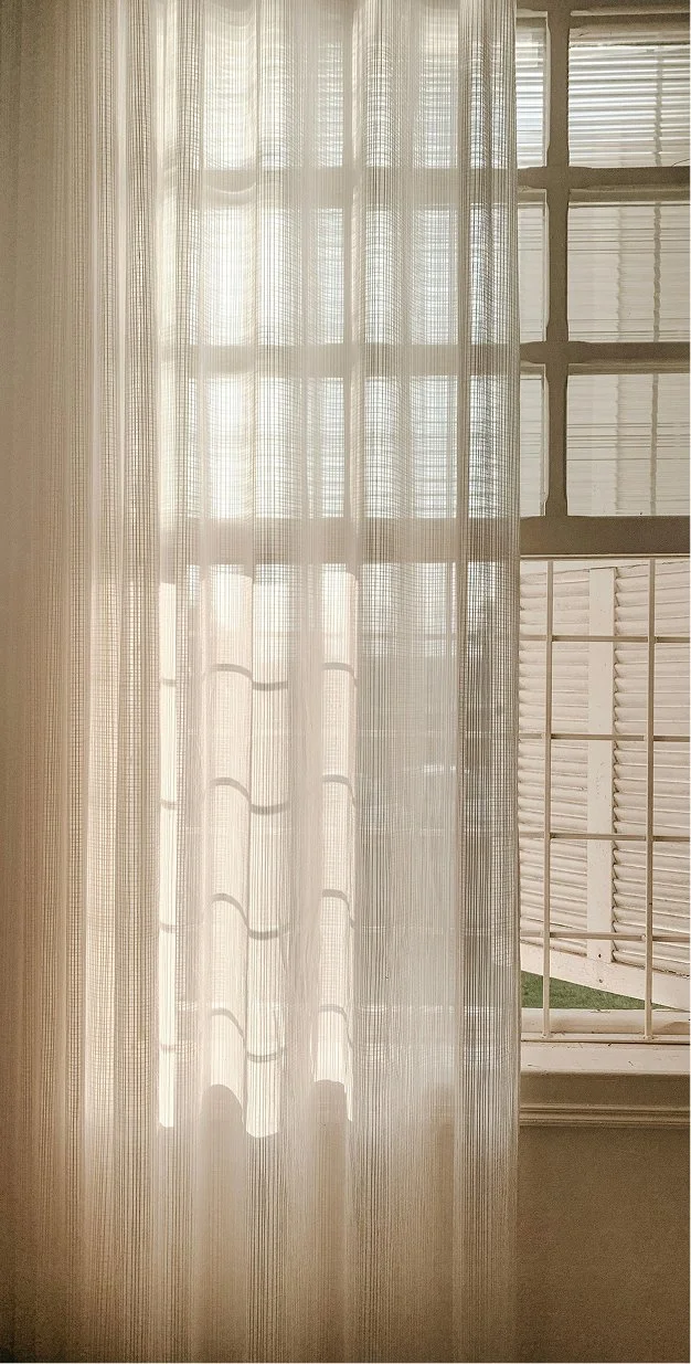 Sunlight shines through sheer curtains onto a window with wooden shutters outside.