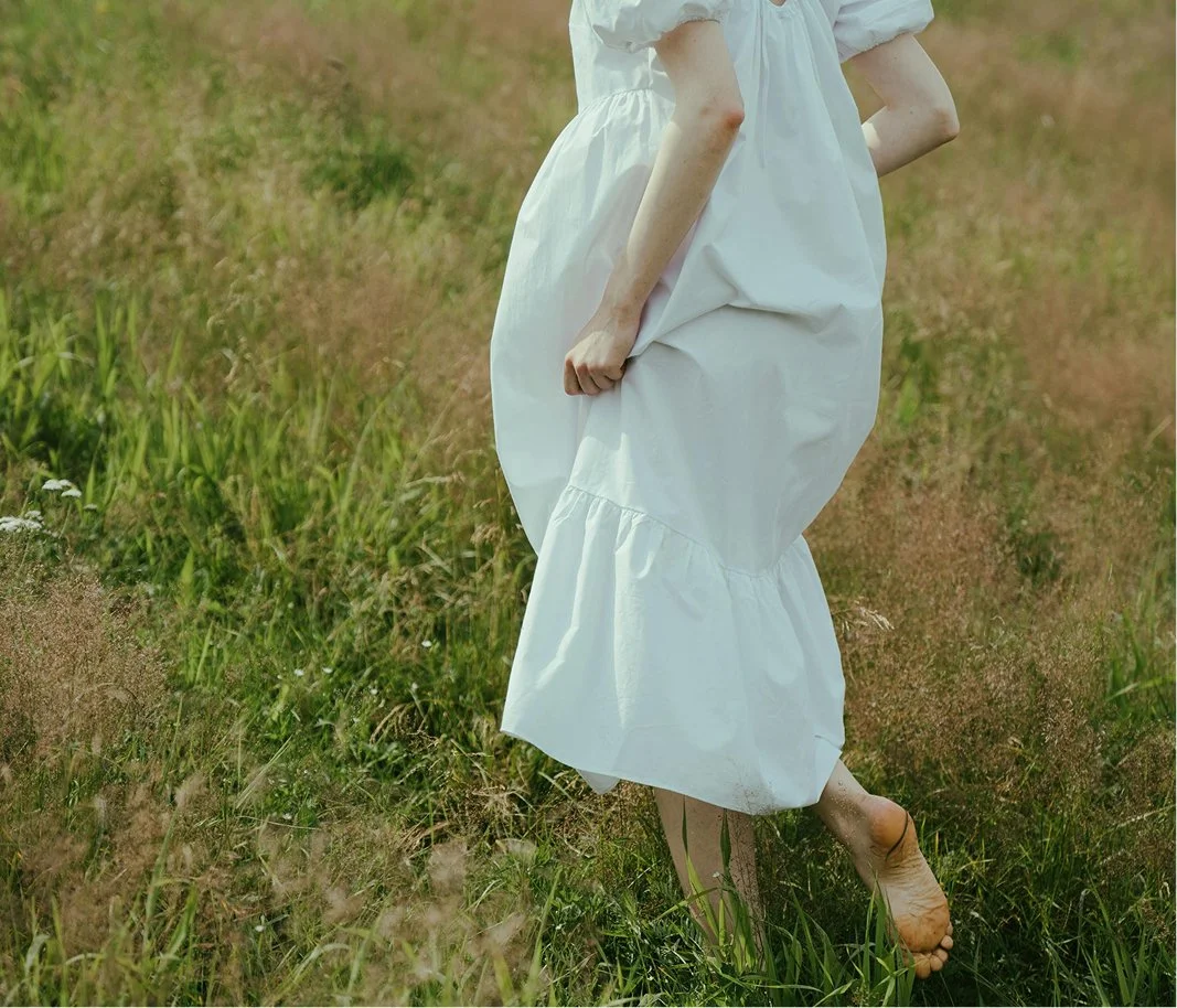 Person wearing a white dress standing barefoot in a grassy field.