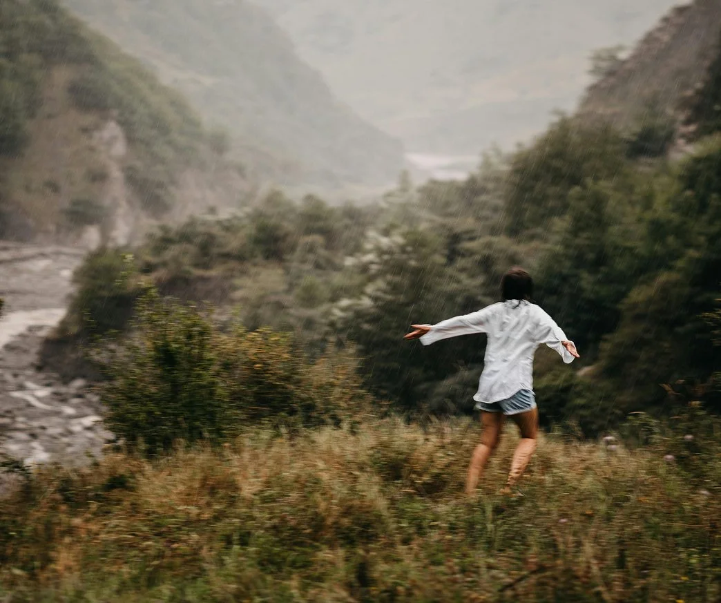Person running through a grassy field in the rain with mountains and trees in the background.