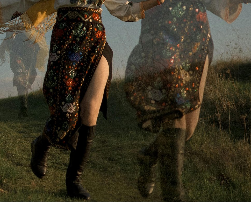 Two people walking through a grassy field, dressed in traditional, colorful, patterned skirts with high slits and knee-high boots, during sunset or twilight.