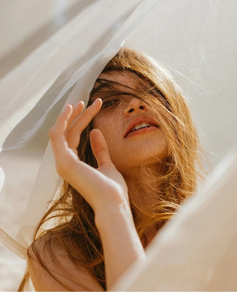 A woman with tousled red hair posing under white fabric, gazing at the camera with her hand near her face.
