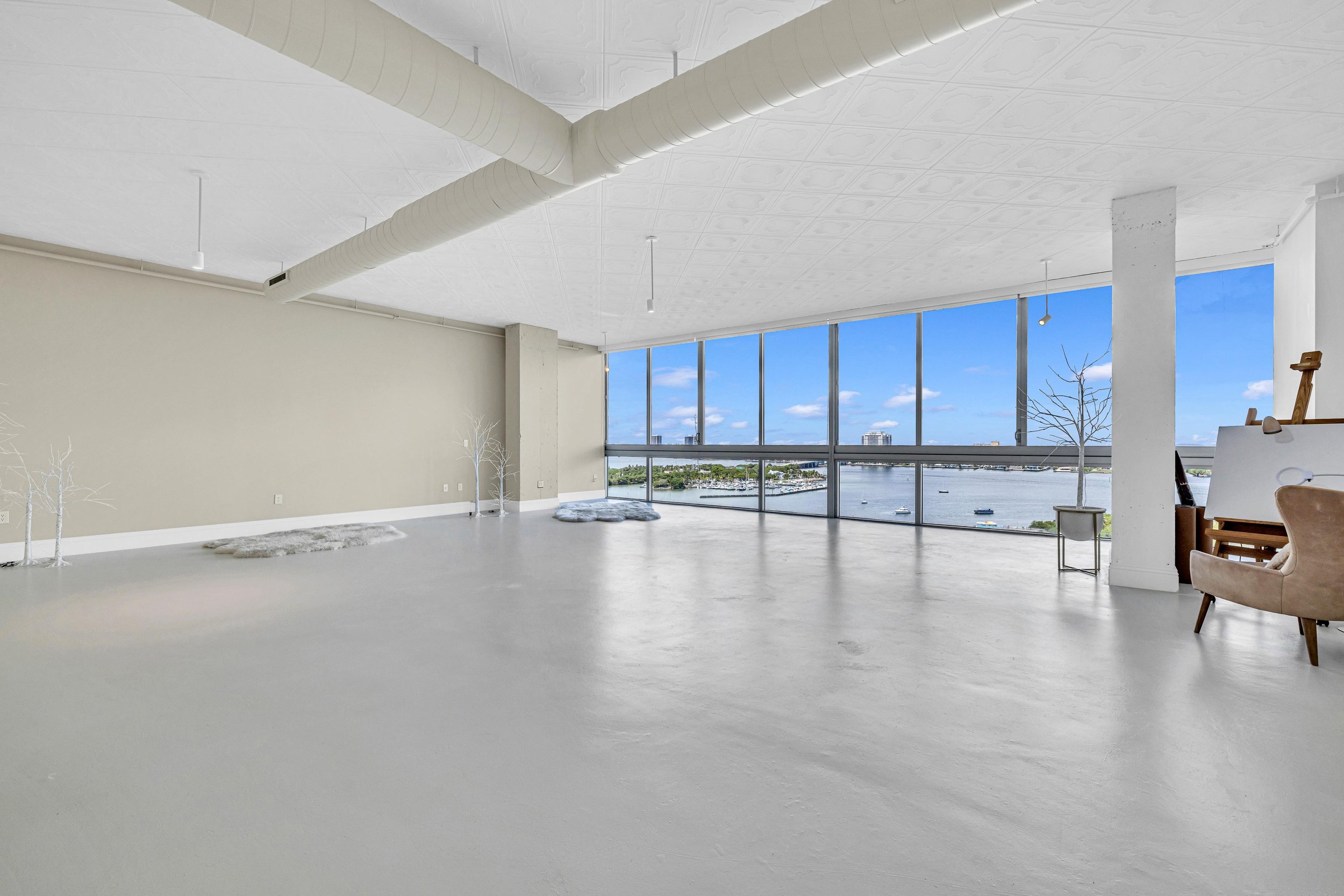 Empty room with large floor-to-ceiling windows overlooking a body of water, minimal decor, white walls, concrete flooring, and some outdoor trees visible through the windows.