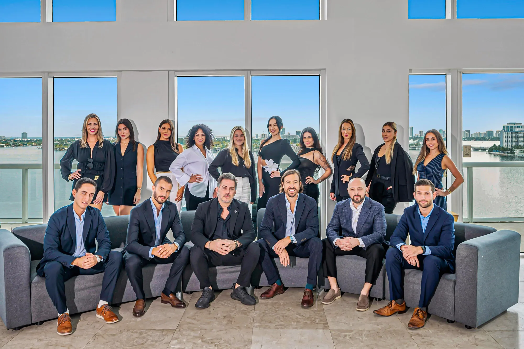 Group of professionals, men and women, in business attire, posing in a modern office with large windows and city view.