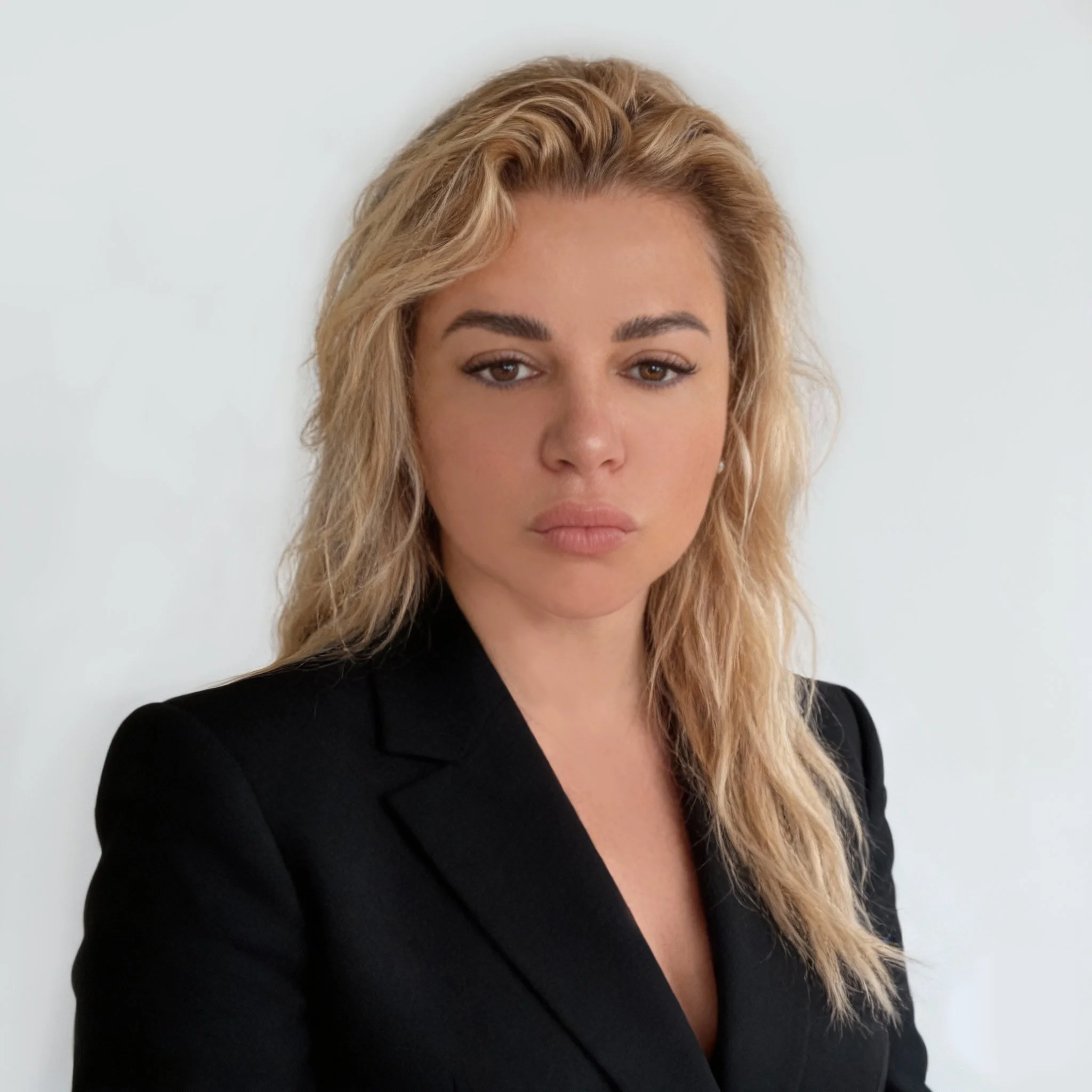 A woman with wavy blonde hair wearing a black blazer against a plain white background.