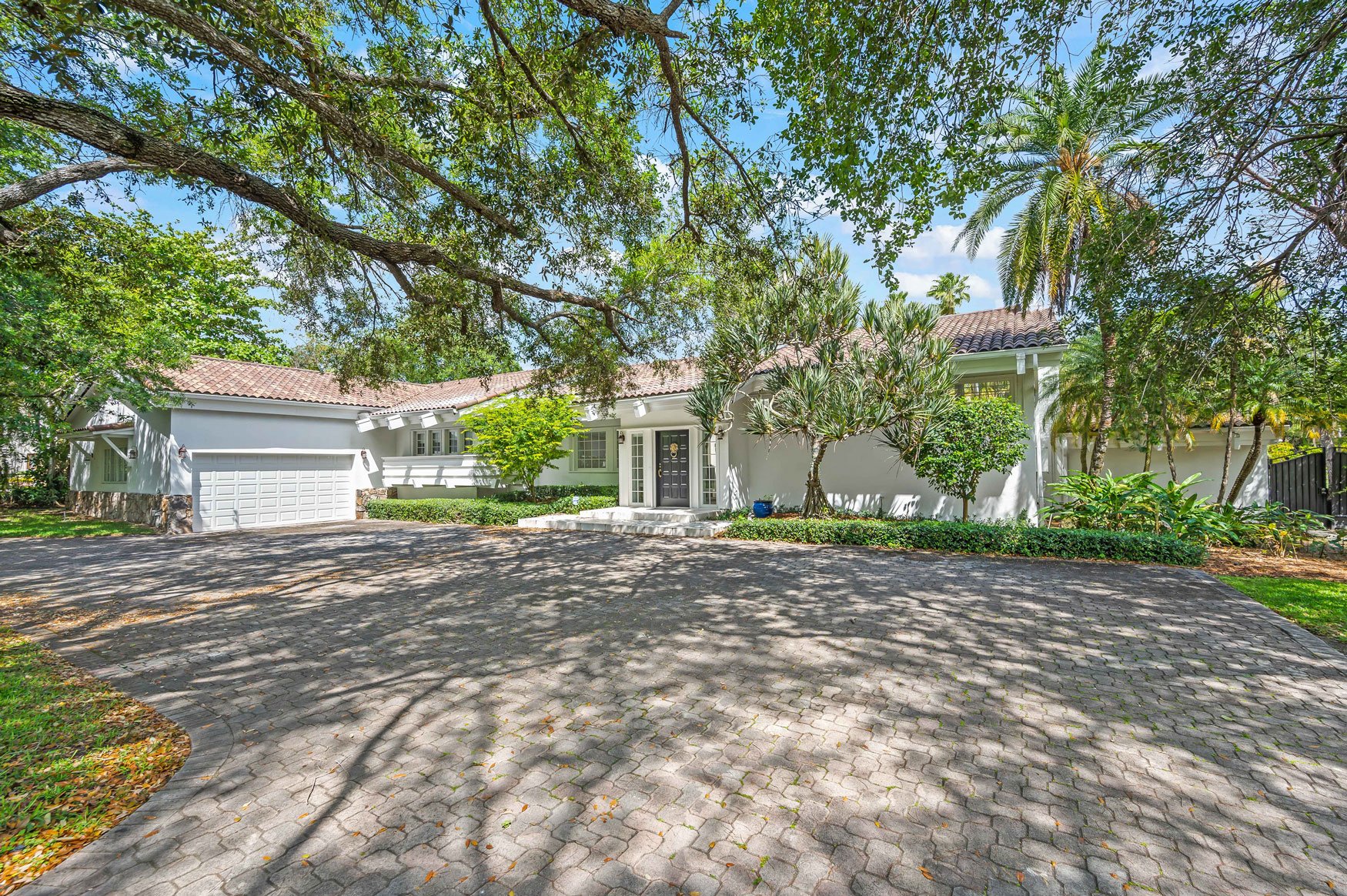 A white house with a stone foundation, double garage, and front steps, surrounded by lush greenery and trees, with a paved driveway.