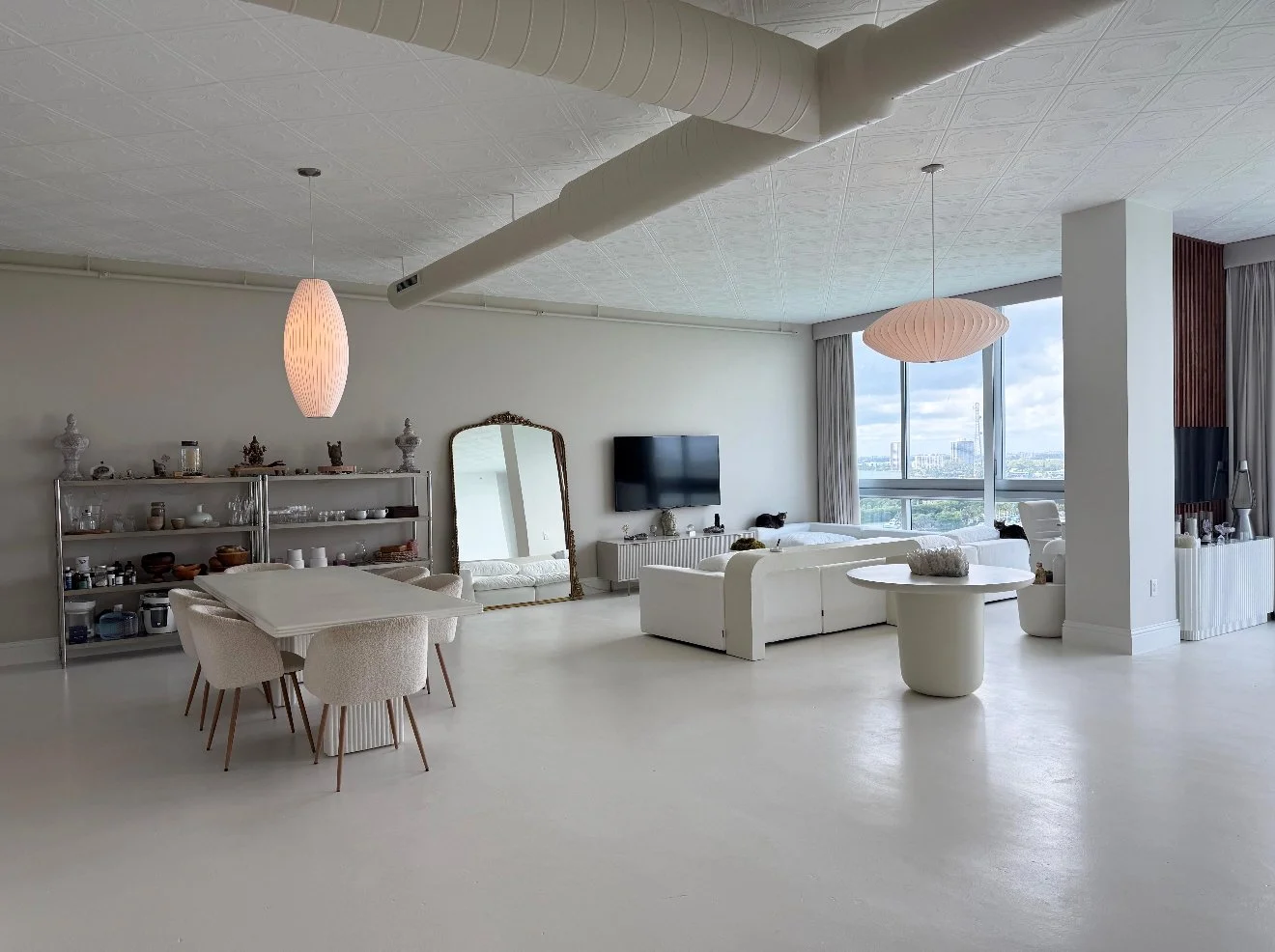 Modern, minimalist living room with white furniture, a dining table with chairs, a large mirror, a TV, and large windows with city view, decorated with pendant lights and decorative objects.