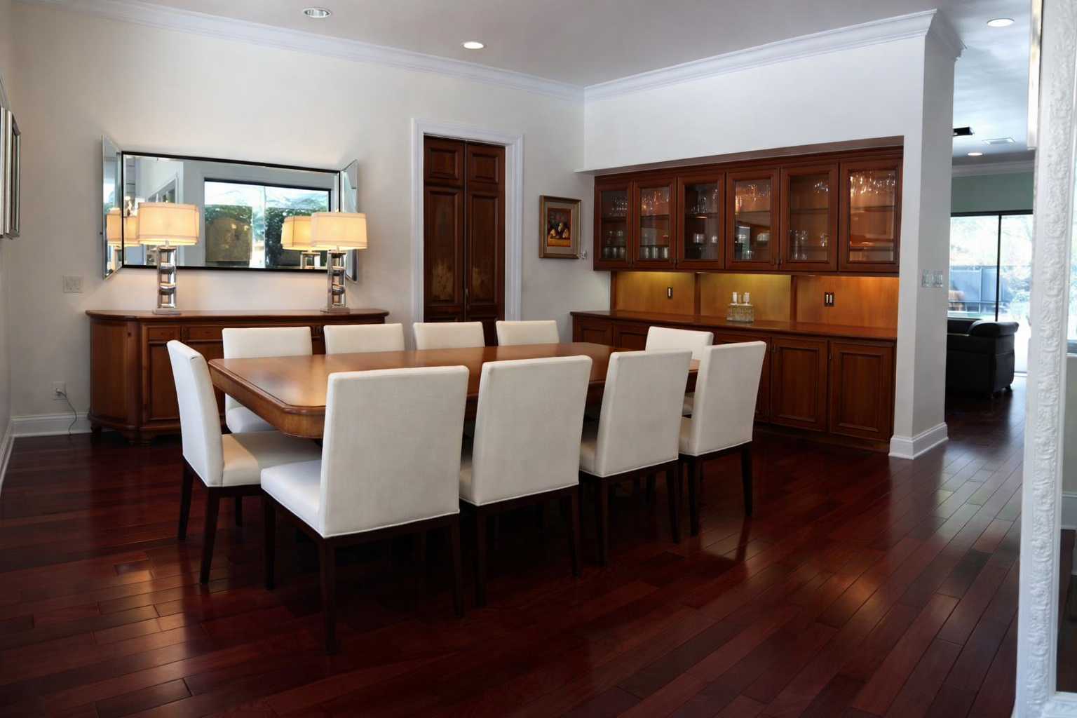 Dining room with a large wooden table surrounded by nine white upholstered chairs, a wooden china cabinet, a sideboard with lamps, a mirror, and a window with green trees outside.