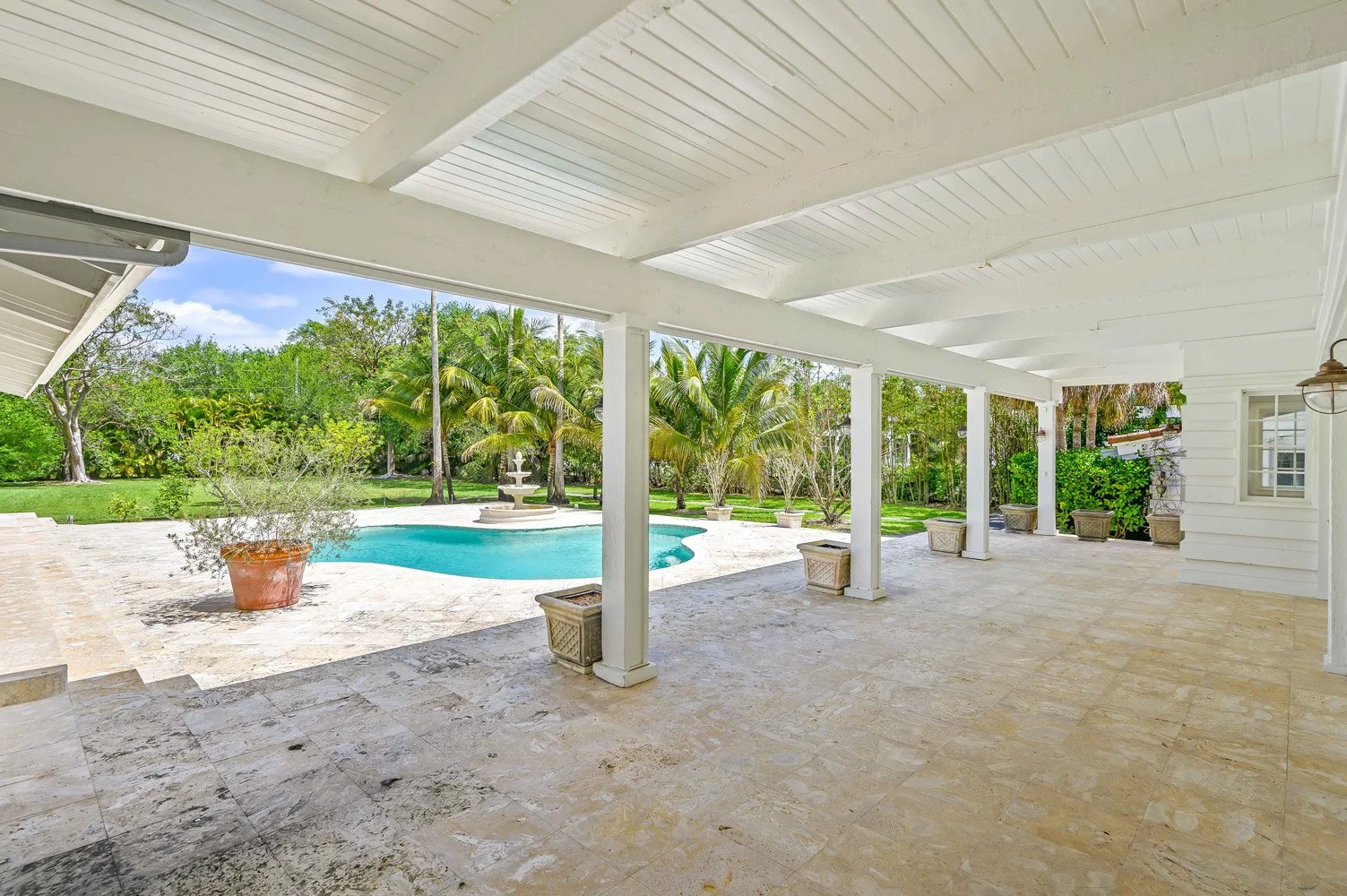 Covered patio area with tiled floor overlooking a backyard with a swimming pool, surrounded by lush greenery and palm trees.