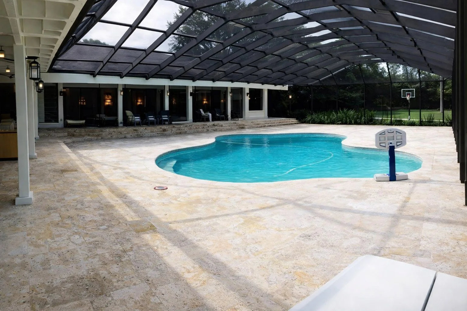 Indoor swimming pool surrounded by beige stone tiles with a basketball hoop at the far end, enclosed by a glass ceiling and walls, with patio chairs along the back wall and greenery outside.