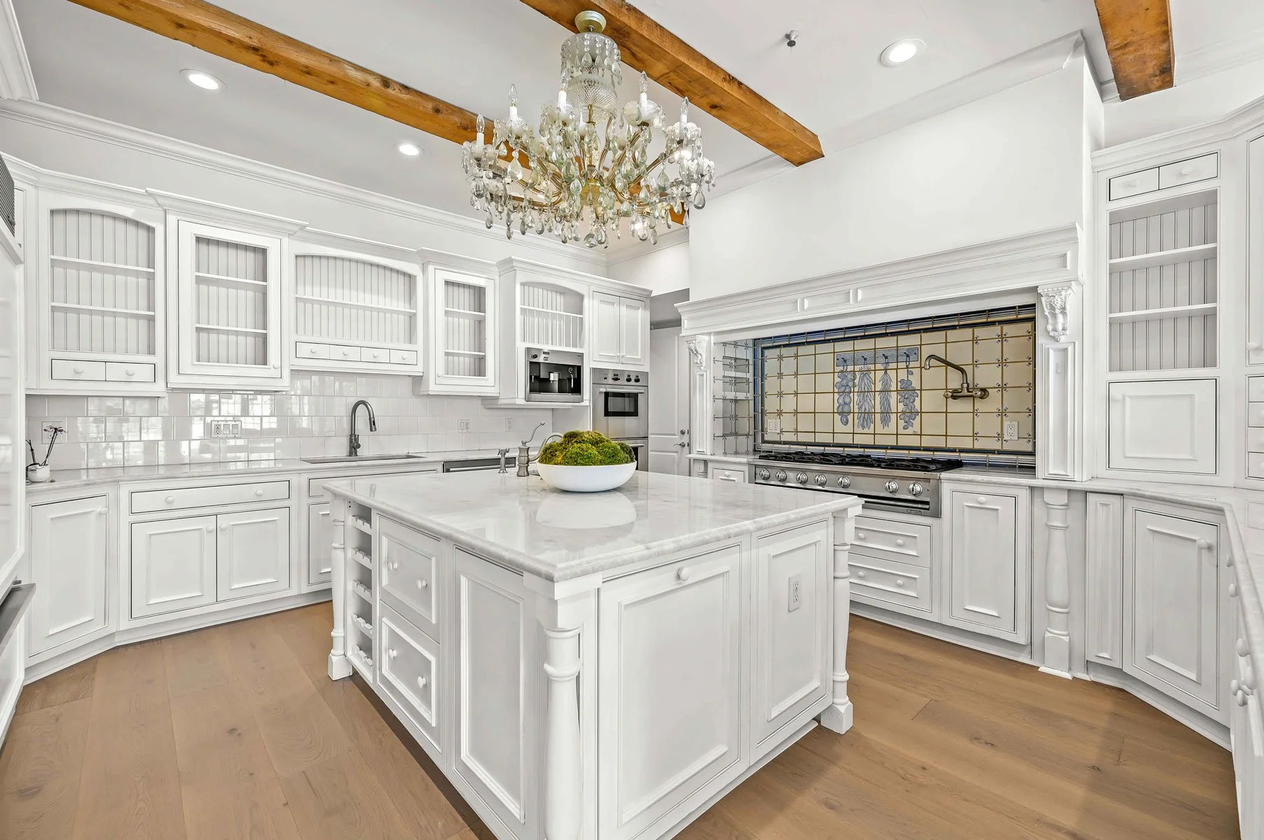 White kitchen with wooden beams, a chandelier, and a marble island, featuring white cabinetry, a tiled backsplash, and stainless steel appliances.