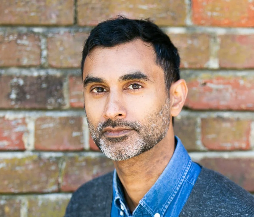 Portrait of a man with dark hair, a beard, wearing a blue collared shirt and a dark sweater, standing in front of a weathered brick wall.
