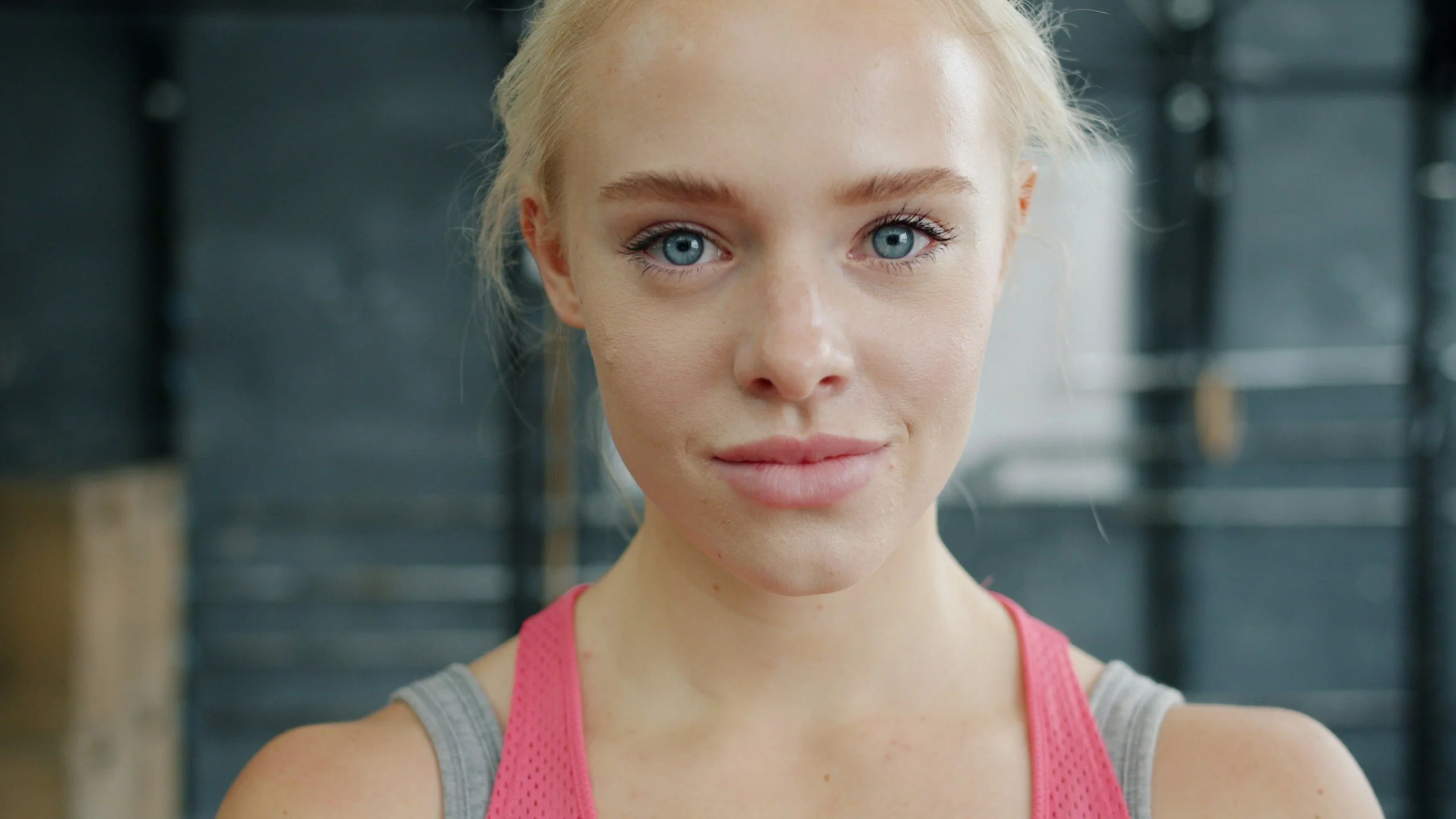 A young woman with blonde hair and blue eyes, wearing a pink sports tank top, in a gym setting.