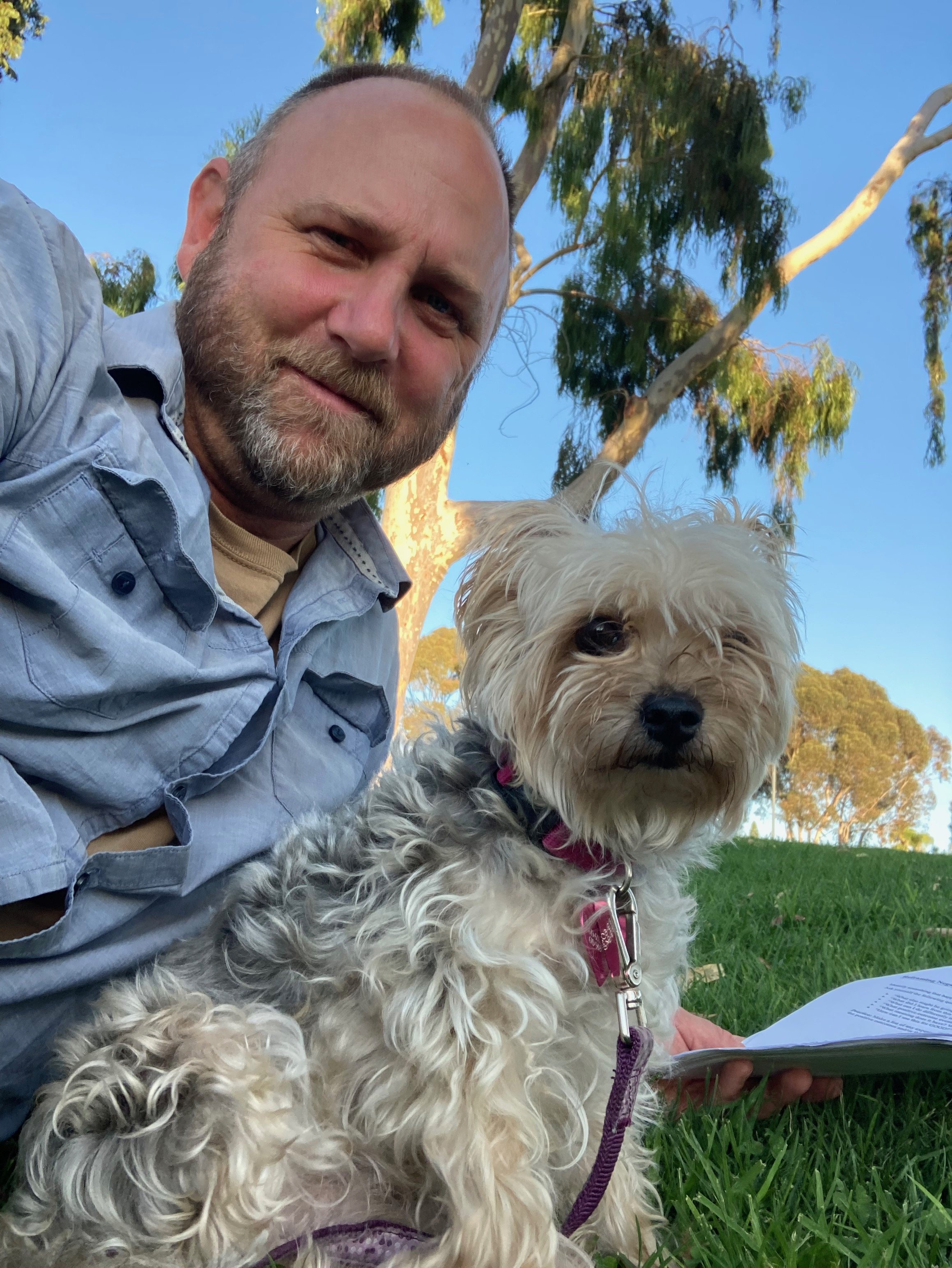 Dog Uncle Nate and a dog sitting together on grass outdoors under trees with a clear blue sky in San Diego.