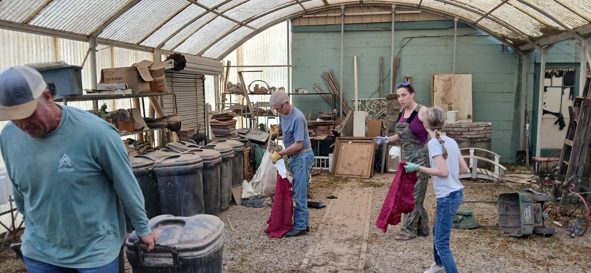 People cleaning and organizing an indoor space filled with clutter, boxes, and various objects, with a curved roof and green walls.